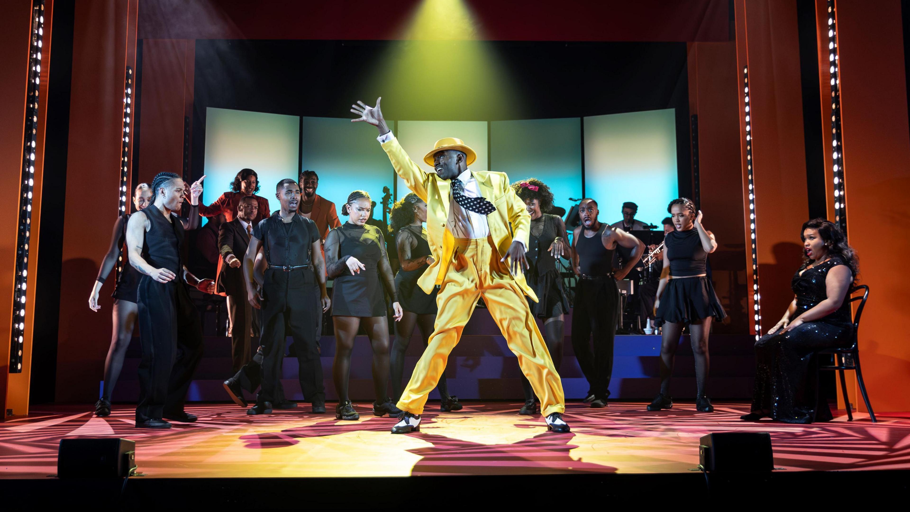 A group of performers stand on stage at the Bristol Old Vic, with one man standing out in front wearing an all-yellow suit. They are in the musical Play On, a show based on Shakespeare's 12th night. Behind the man at the front are dozens of performers mostly wearing black