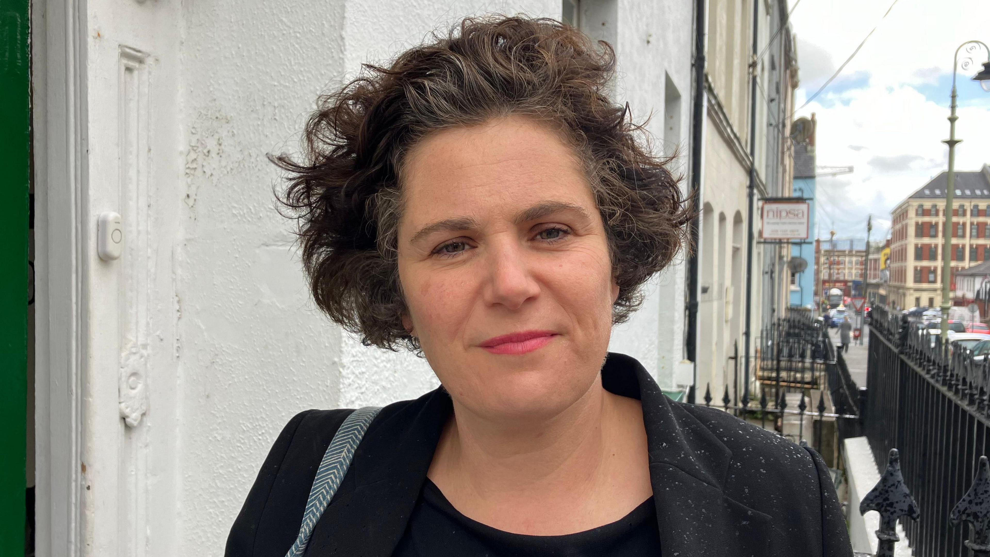 Claire Hanna on a busy street wearing a dark suit. Behind her is a white wall, and a green door. To her right are rows of black railings 