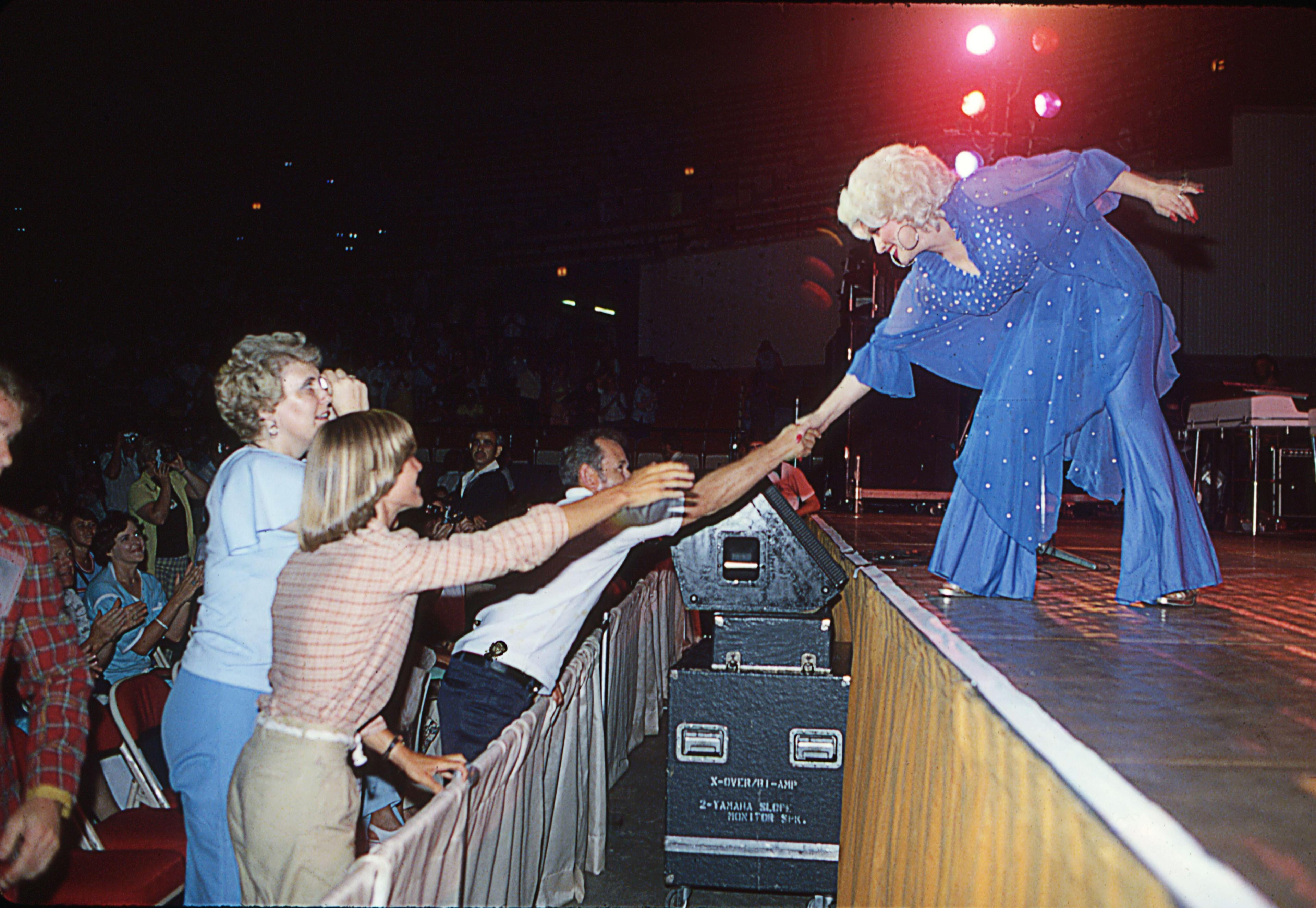 At a live concert, country music superstar Dolly Parton goes to the front of the stage to shake hands with fans. She wears a purple and bedazzled flowy top and pants.