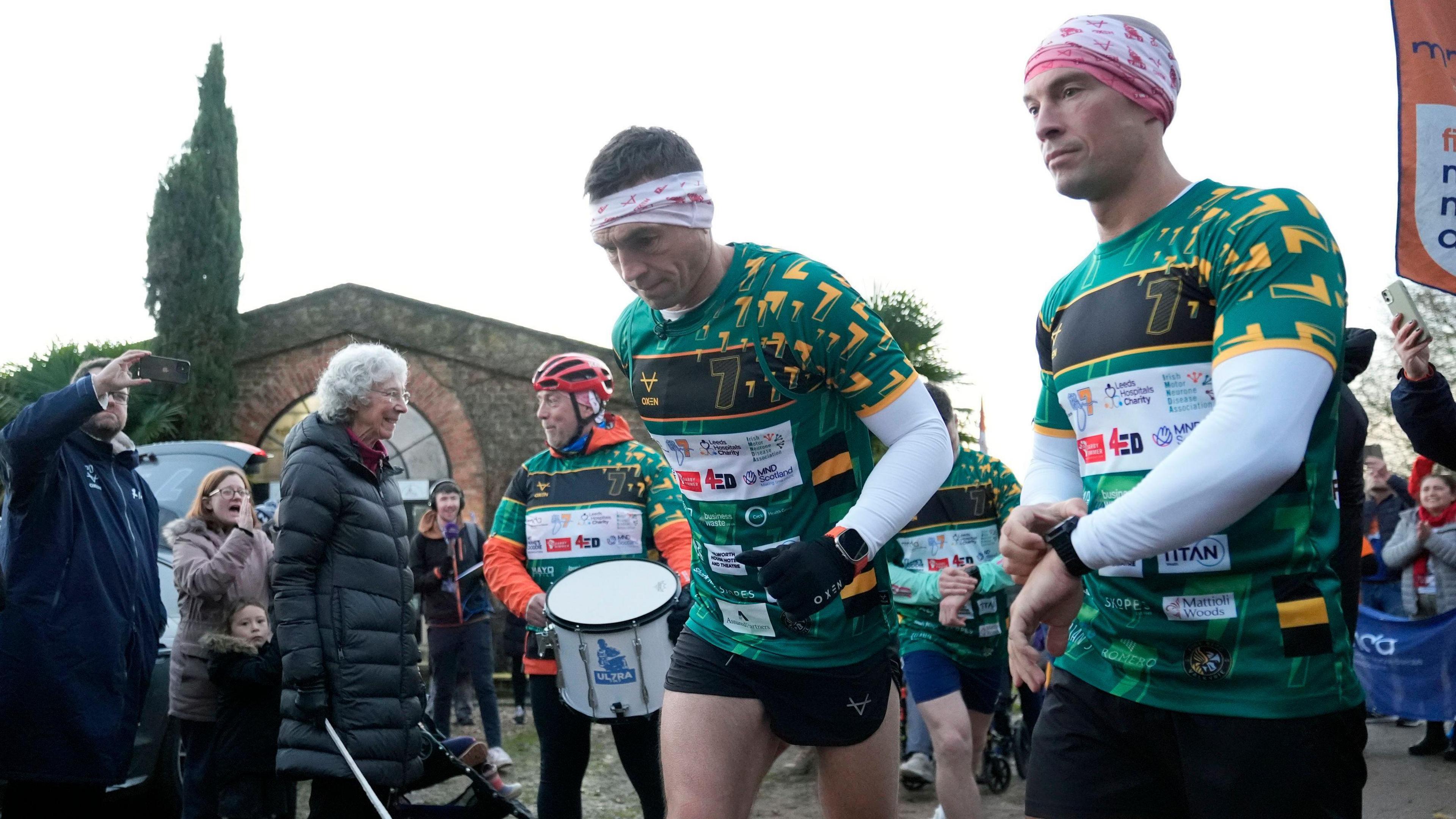 Two men in green and black rugby tops and black shorts and leggings jog past a small crowd of people cheering and clapping them. 