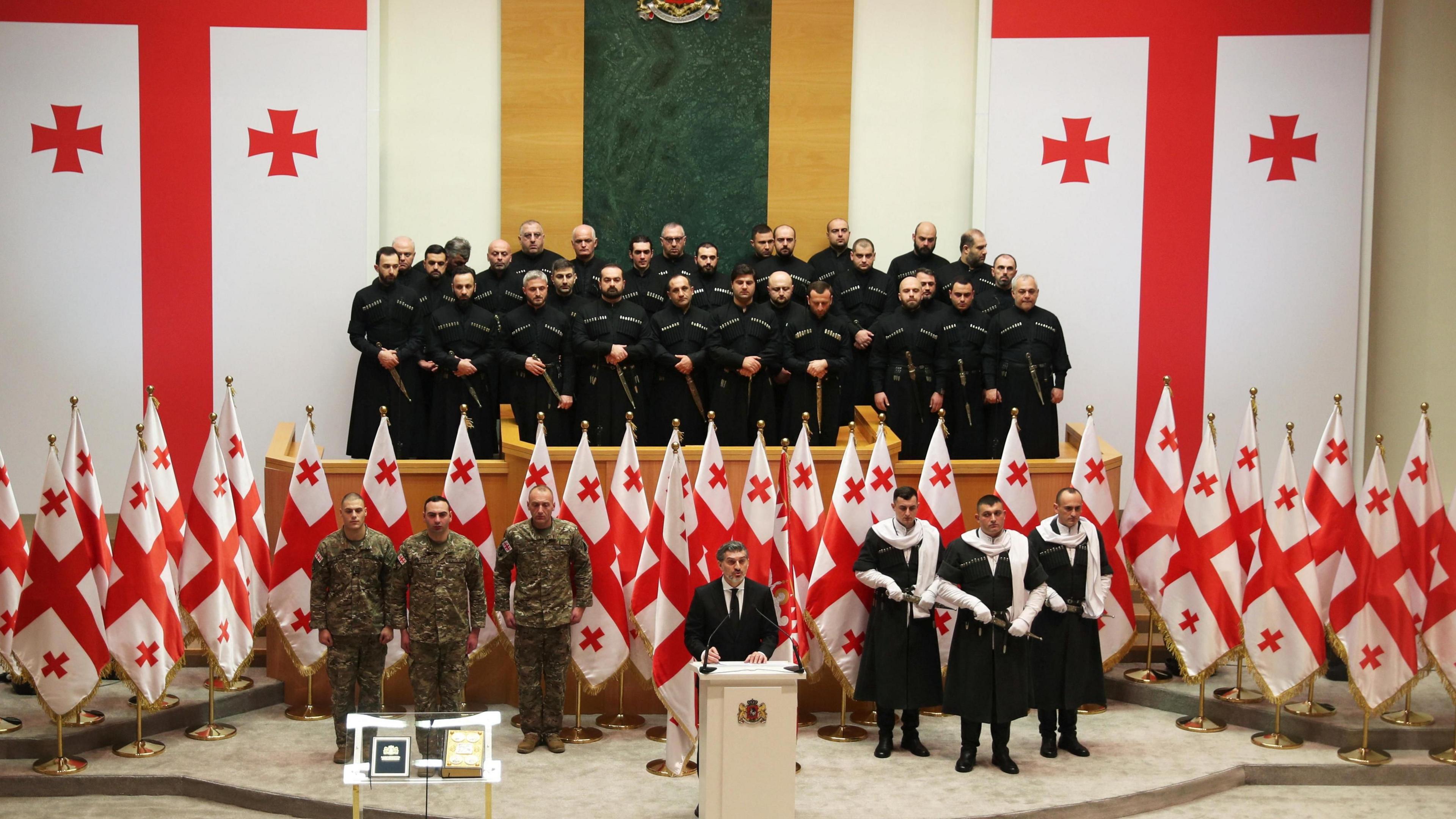 Mikheil Kavelashvili speaking in parliament at is inauguration ceremony, where he is standing in front of Georgian flags and a group of officers with guns