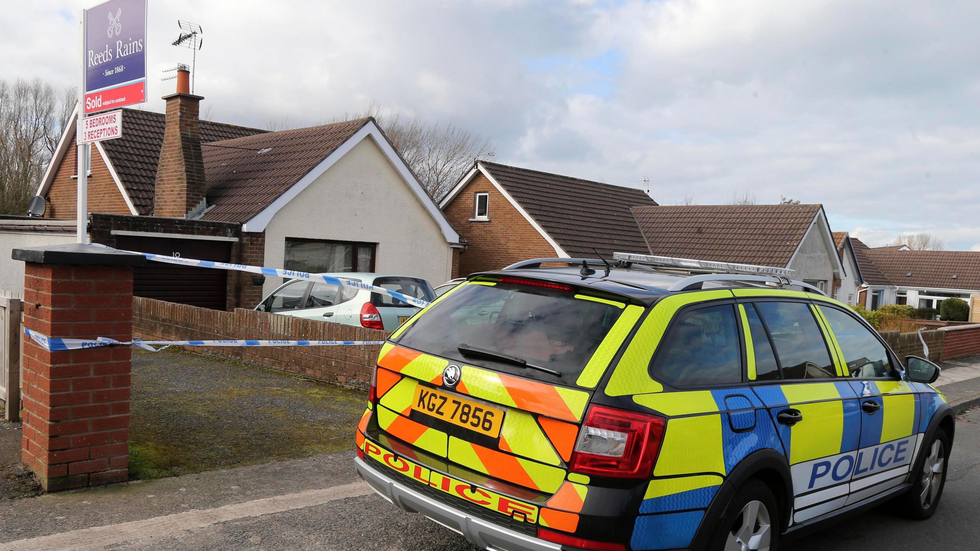 A row of residential homes with a yellow, blue and orange police car on the kerb outside. there is blue and white police tape and a 'sold' house sign. The houses are small bungalows with red bricks, brown roofs and cream paint.