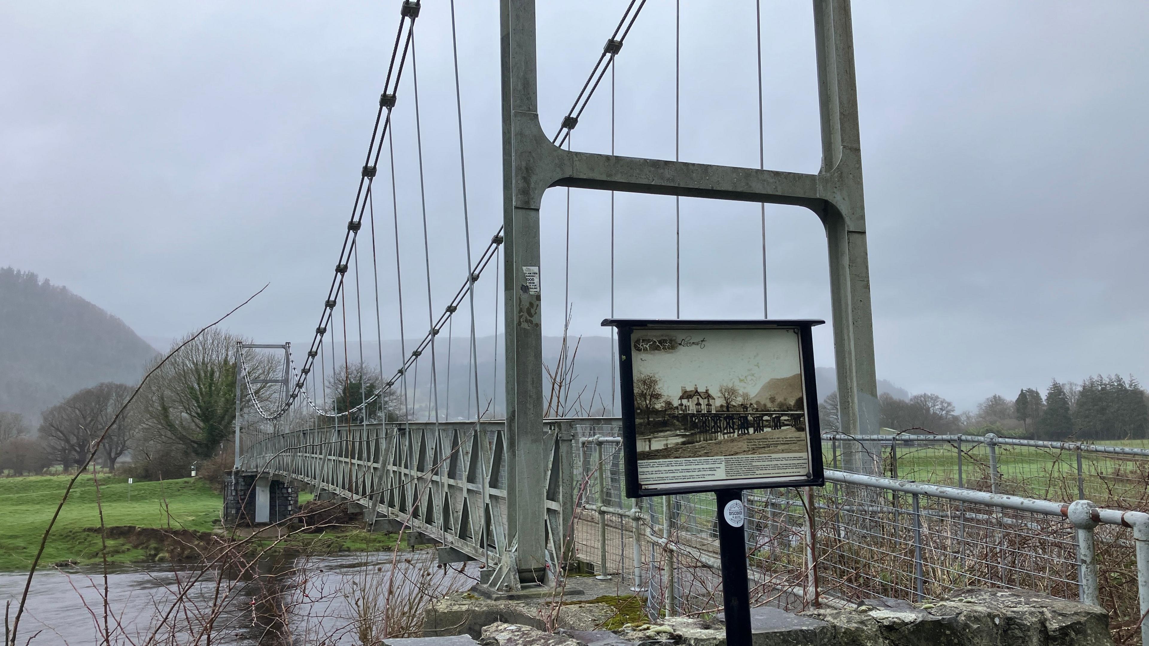 Gower Bridge is seen with a sign explaining its historical significance. In the background there are fields and trees on a foggy day in Llanrwst.