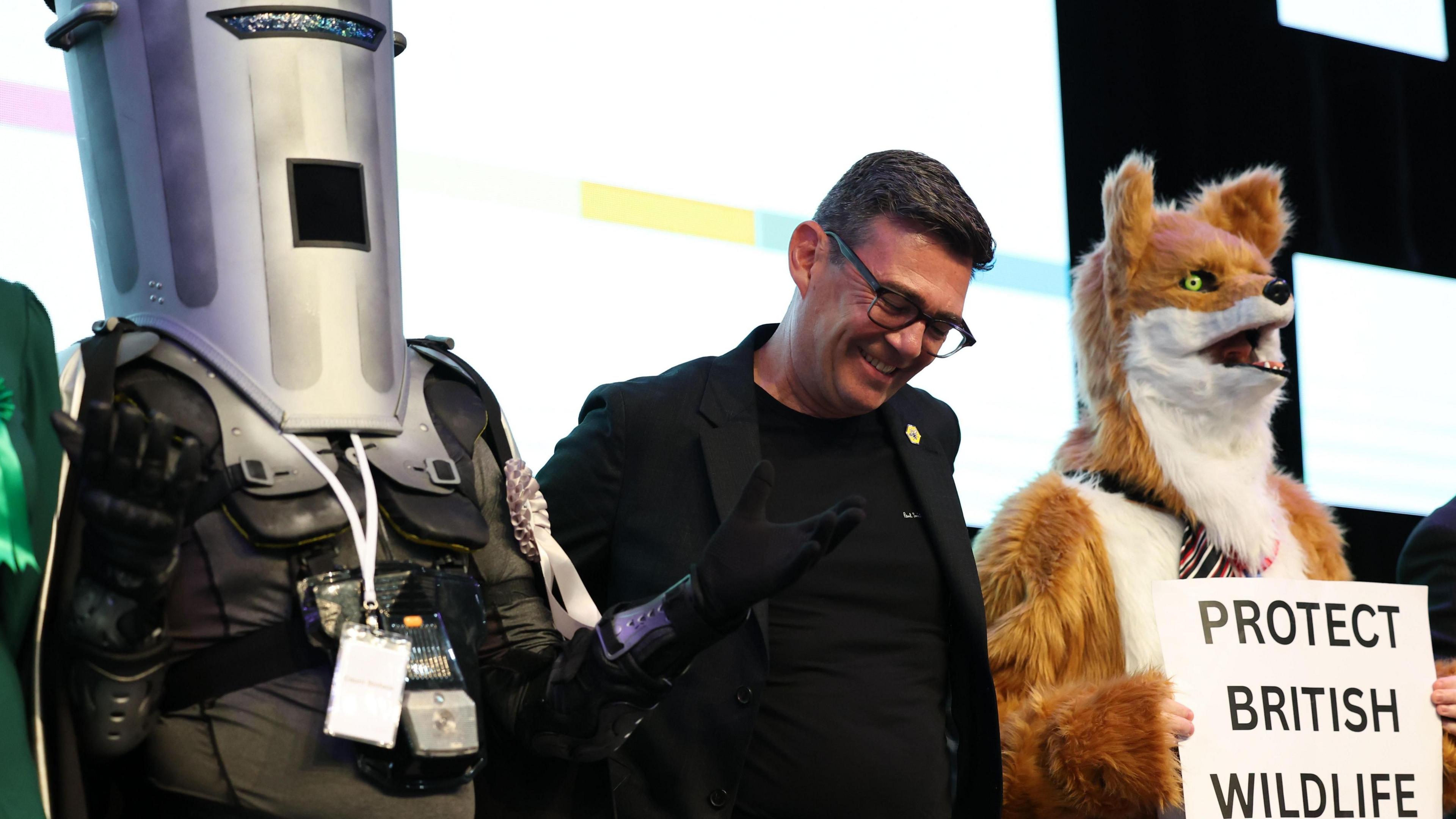 A smiling Andy Burnham stands alongside Count Binface while standing beside a costumed fox after being announced the winner of the Makerfield election.