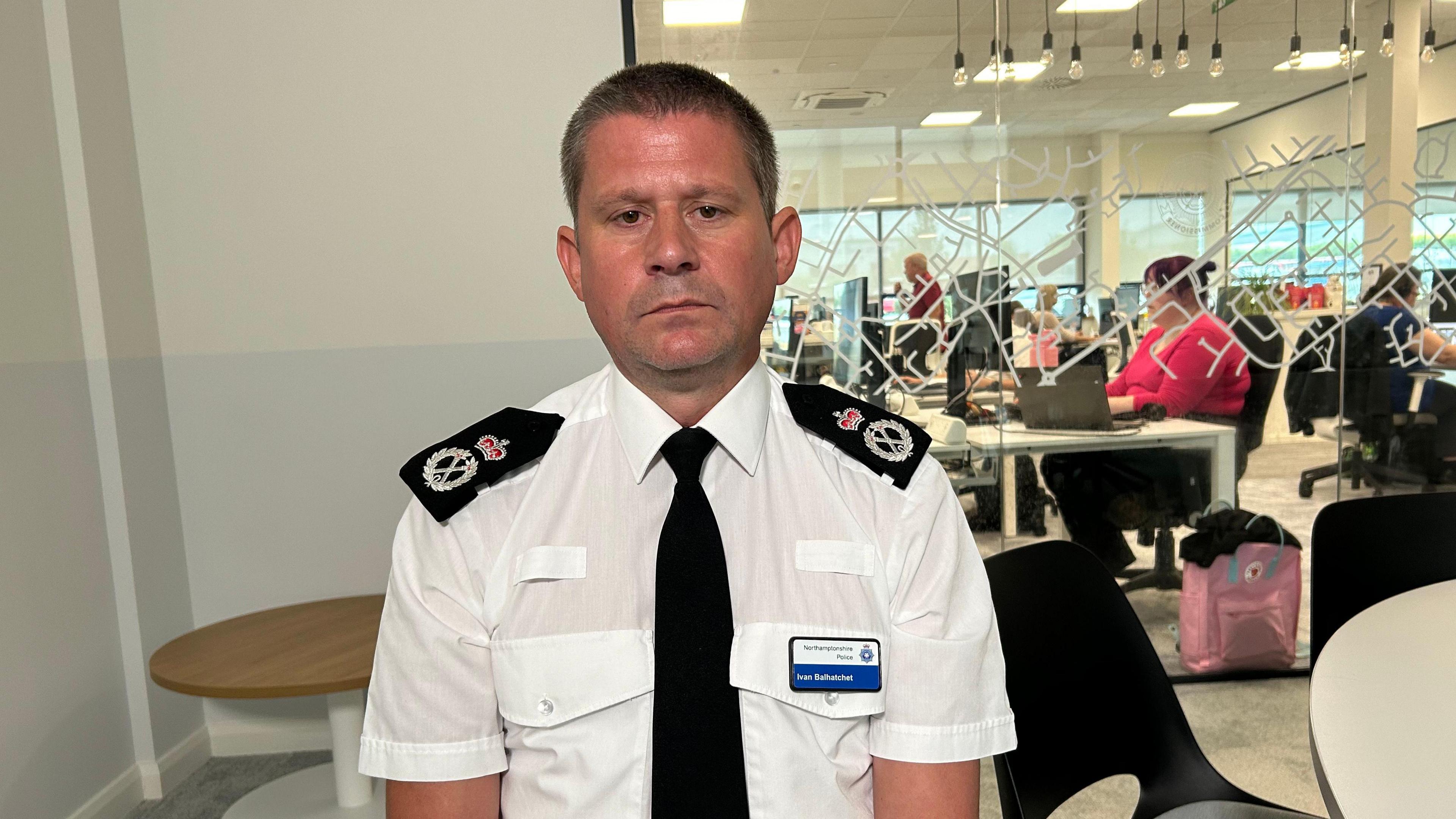 Ivan Balhatchet in police uniform with a white shirt, black tie and epaulettes sits on a chair in an office.