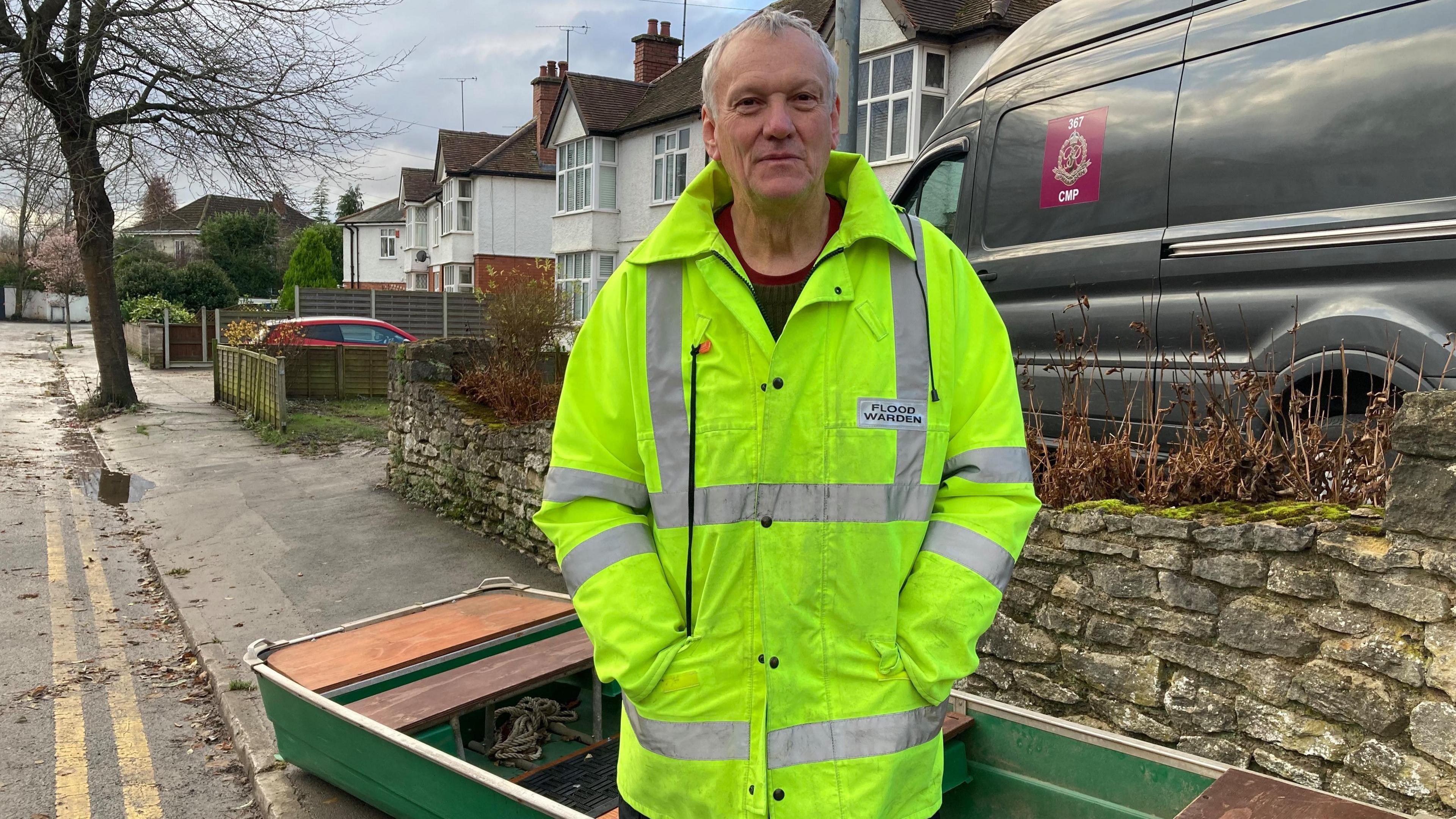 A man with short grey hair stands on a street in front of a small wooden boat. He is wearing a high-vis coat with "FLOOD WARDEN" emblazoned on it.