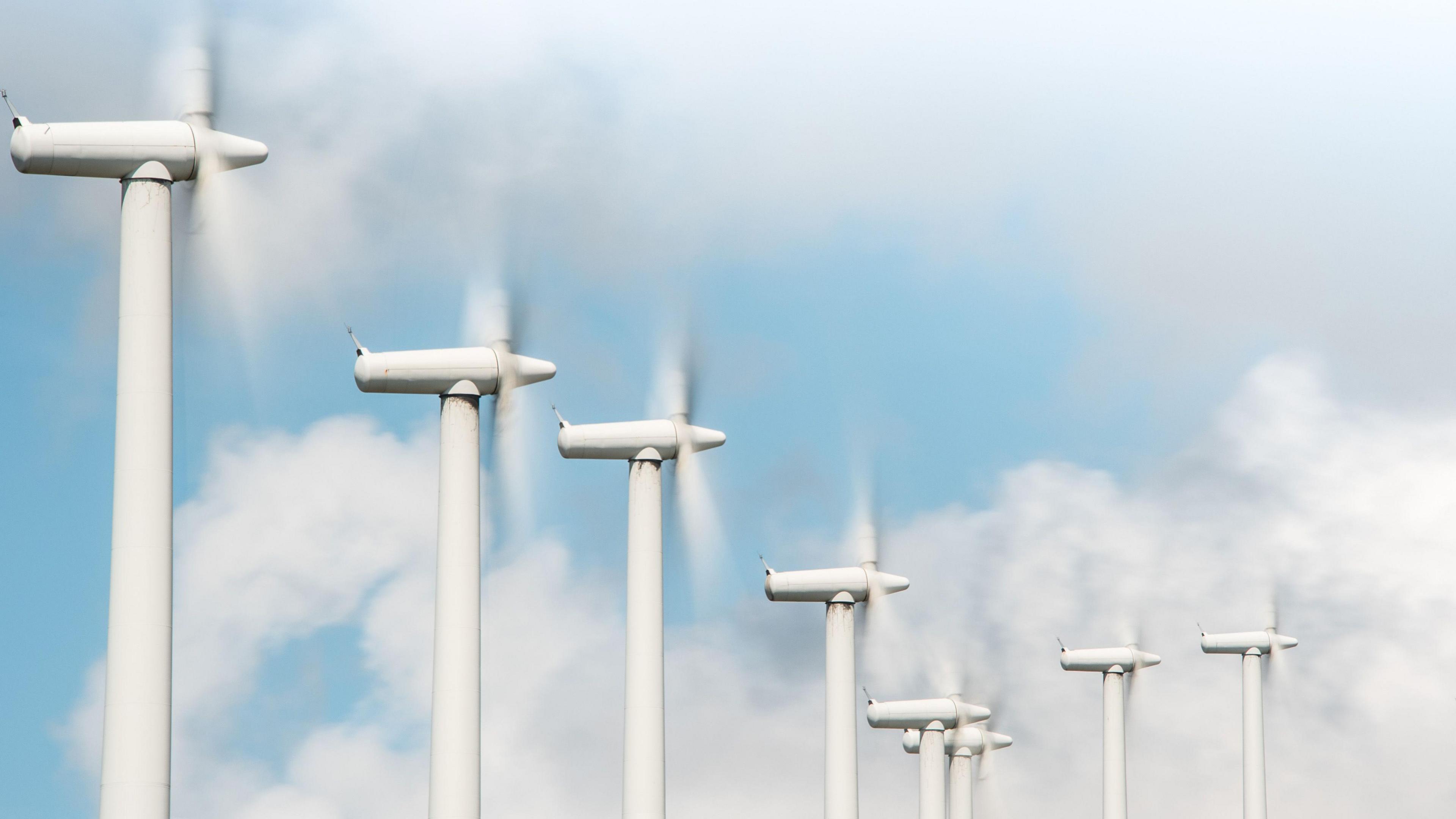 A row of white wind turbines rotating, pictured in front of a blue sky with fluffy white clouds. 