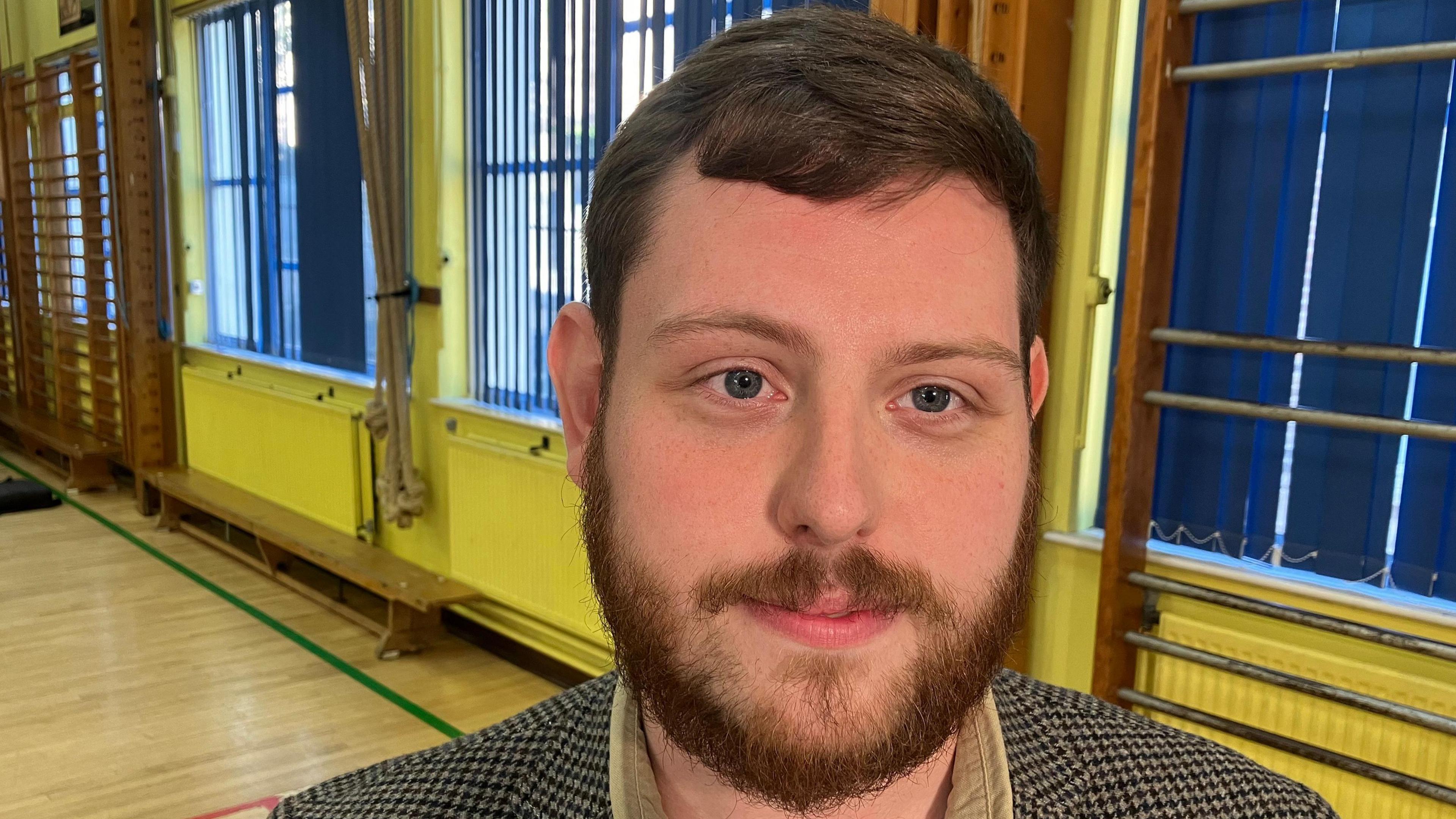 Dylan Sloan who has brown short hair and a beard. He is wearing a plaid suit that's cream and brown with a beige shirt. He is standing in a school hall.