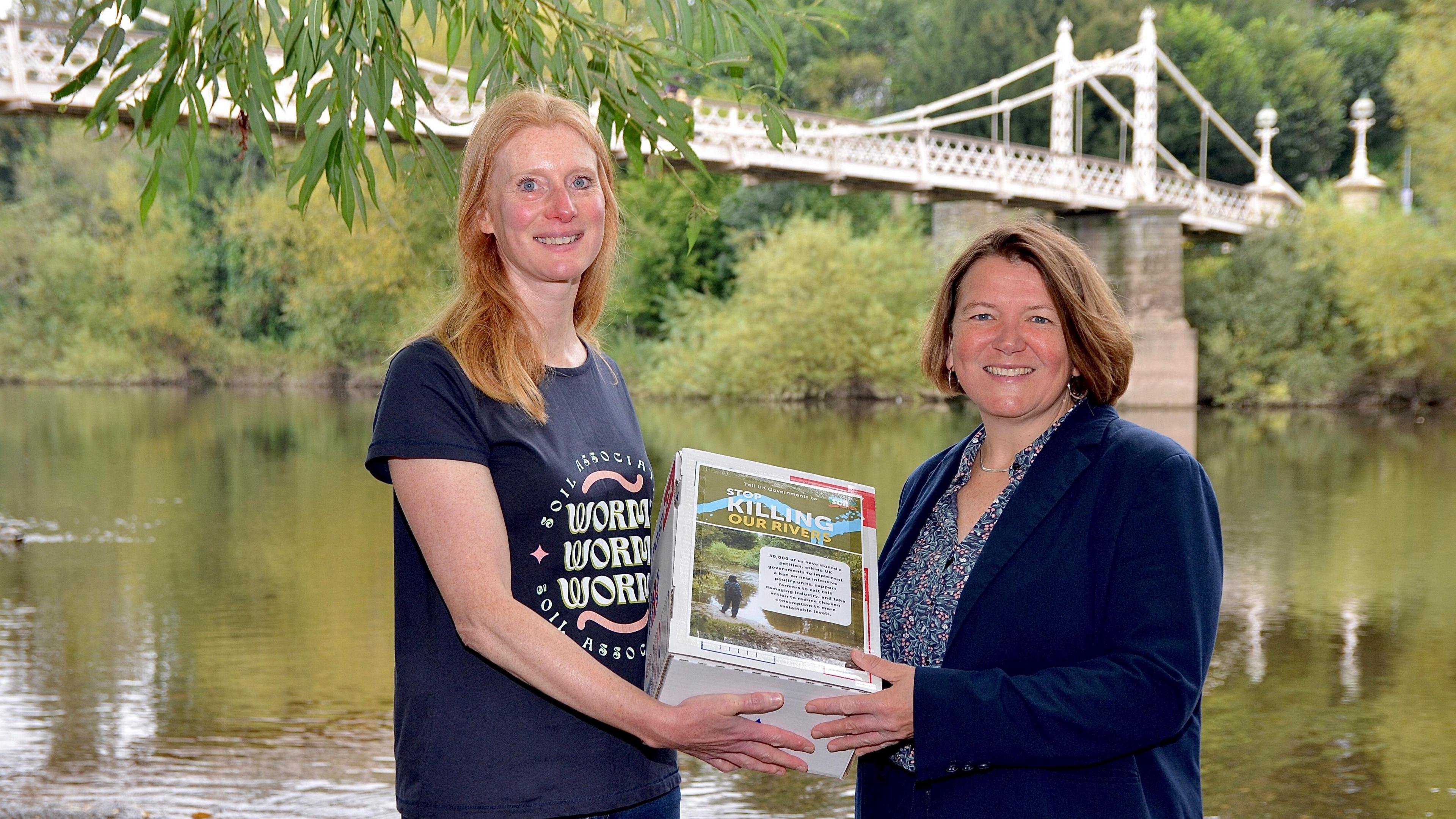 A woman in a navy t-shirt presents a box with a graphic that says Stop Killing Our Rivers to a woman wearing a navy blazer and floral blouse. They are standing in front of a river, in the background a line of trees and a bridge are visible