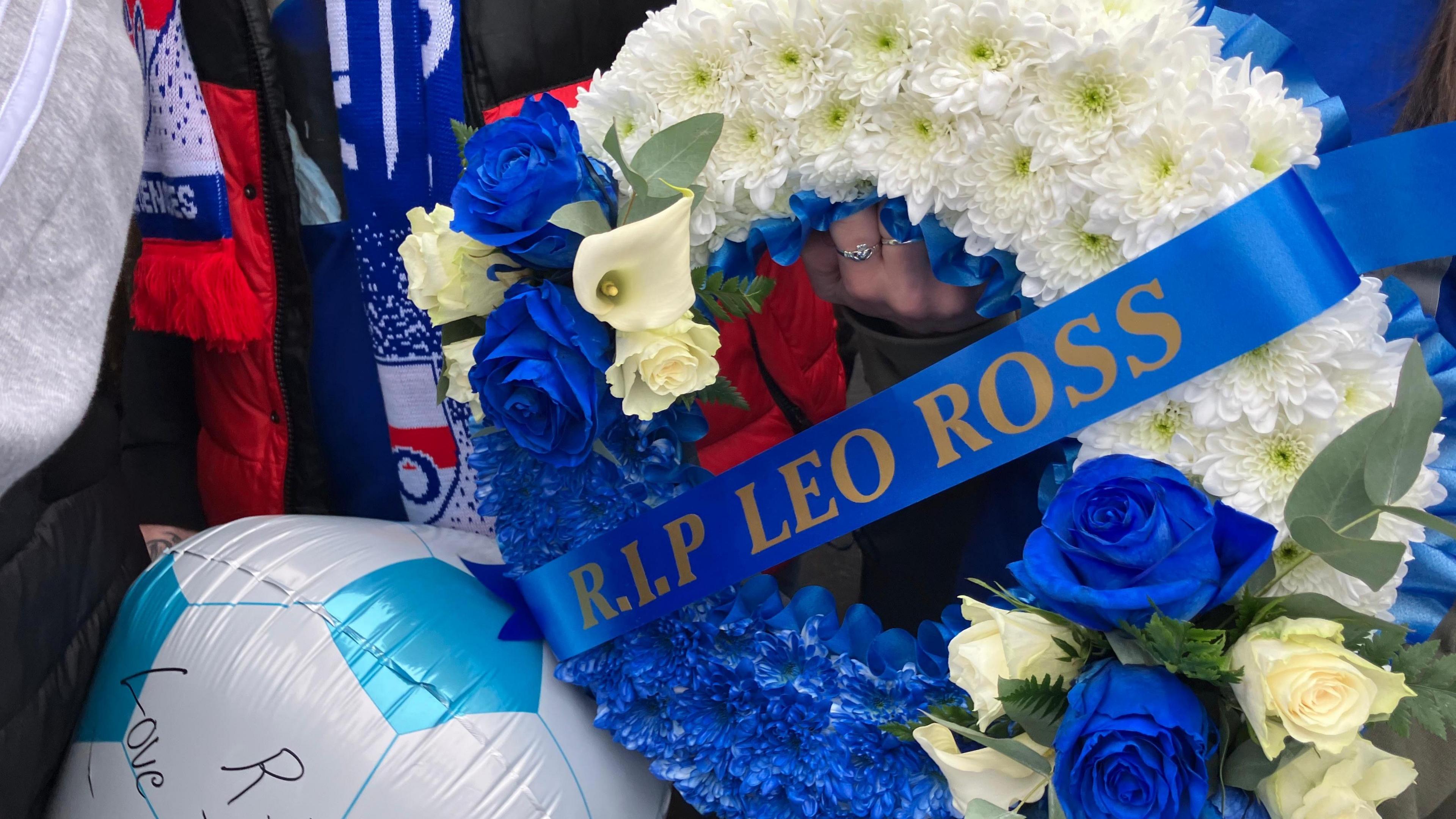 A wreath which reads RIP LEO ROSS. A ringed finger is holding it. Football scarves can be seen in the background. A balloon is also cropped. 