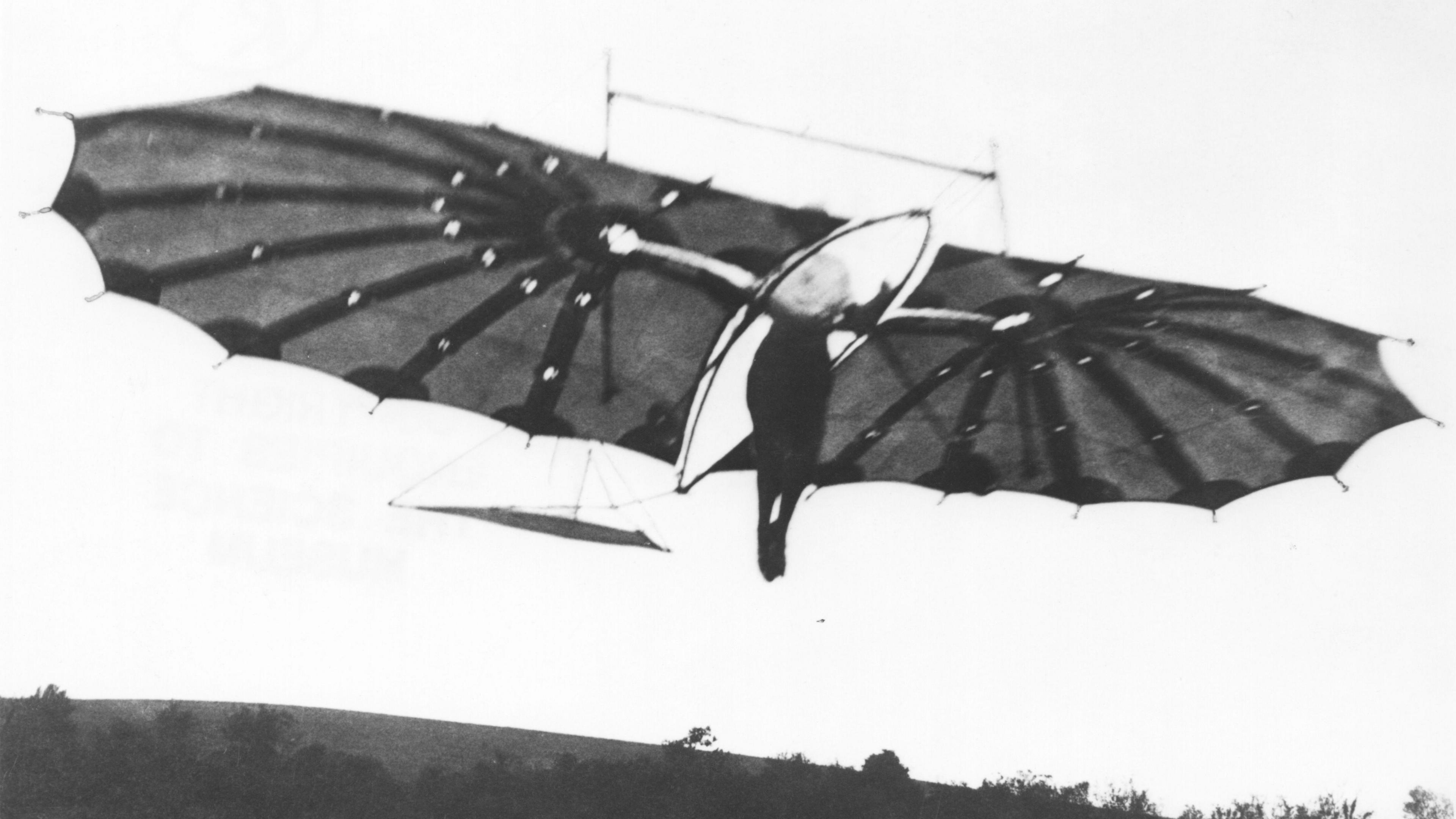 A black and white image showing the Hawk glider in flight. A person dressed in dark clothing can be seen between the wings of the glider.