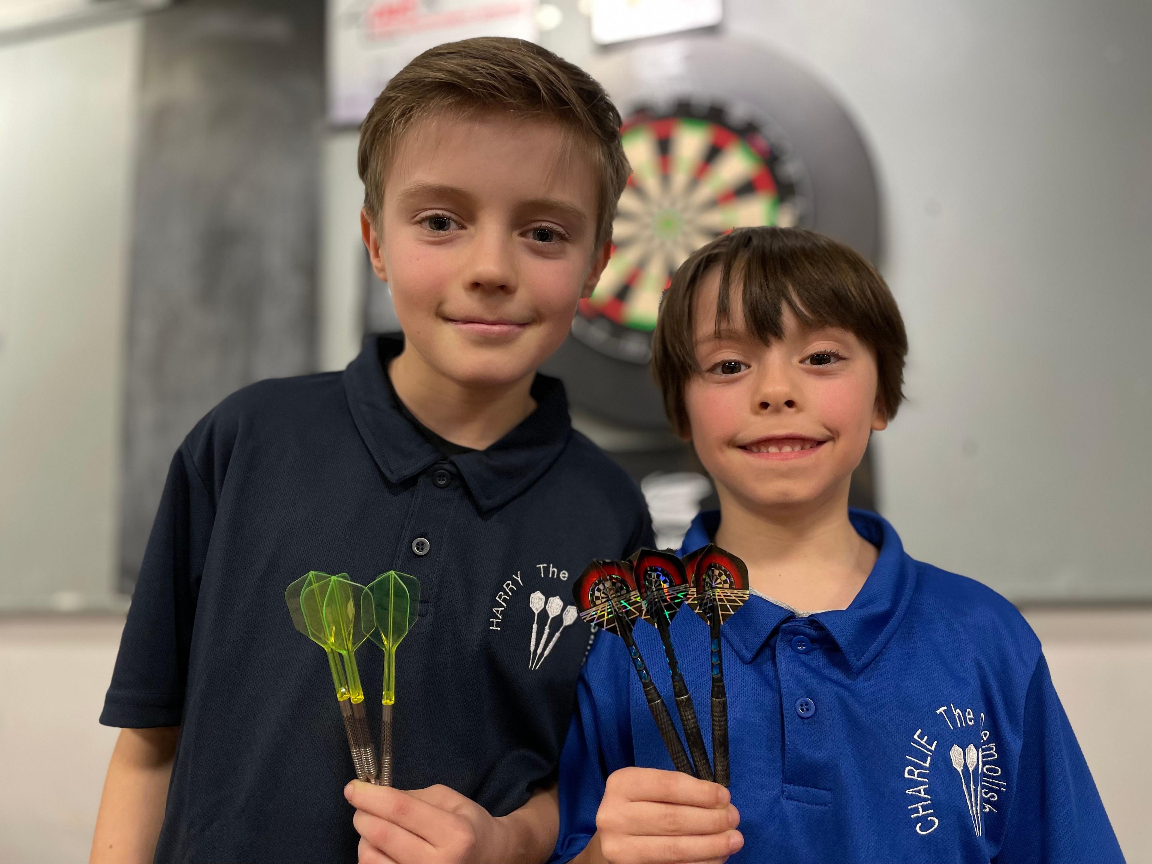 Two boys, Harry, aged 11, and Charlie, aged nine, stand in front of a darts board. They are smiling at the camera. Harry, on the left, is holding a set of three darts in his left hand. The darts have green see-through flights. Charlie, on the right, is holding a set of three darts. The flights are designed with a picture of a darts board.