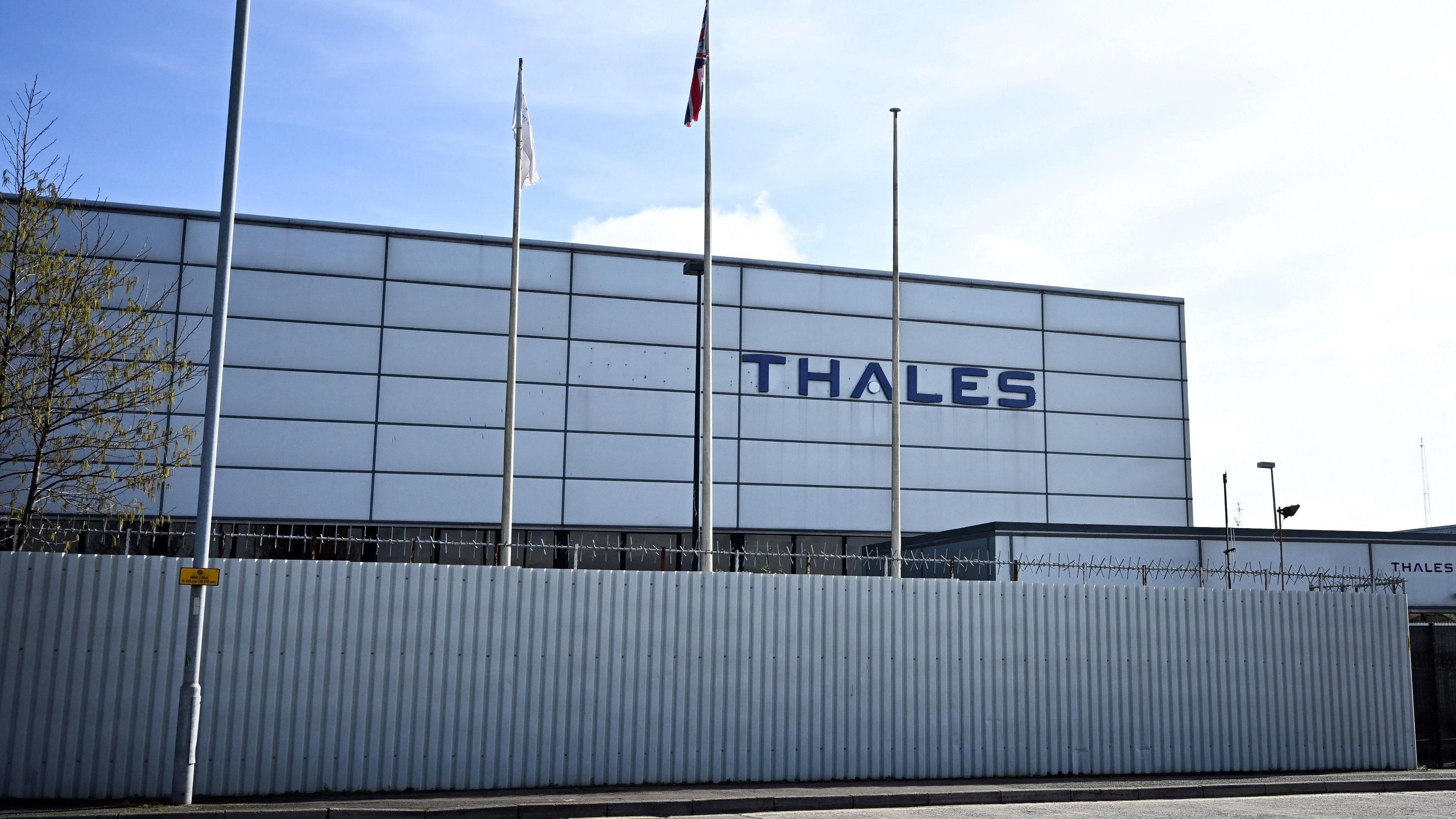 The THALES factory in Belfast. A white corrugated iron building with THALES in blue on the building wall. 