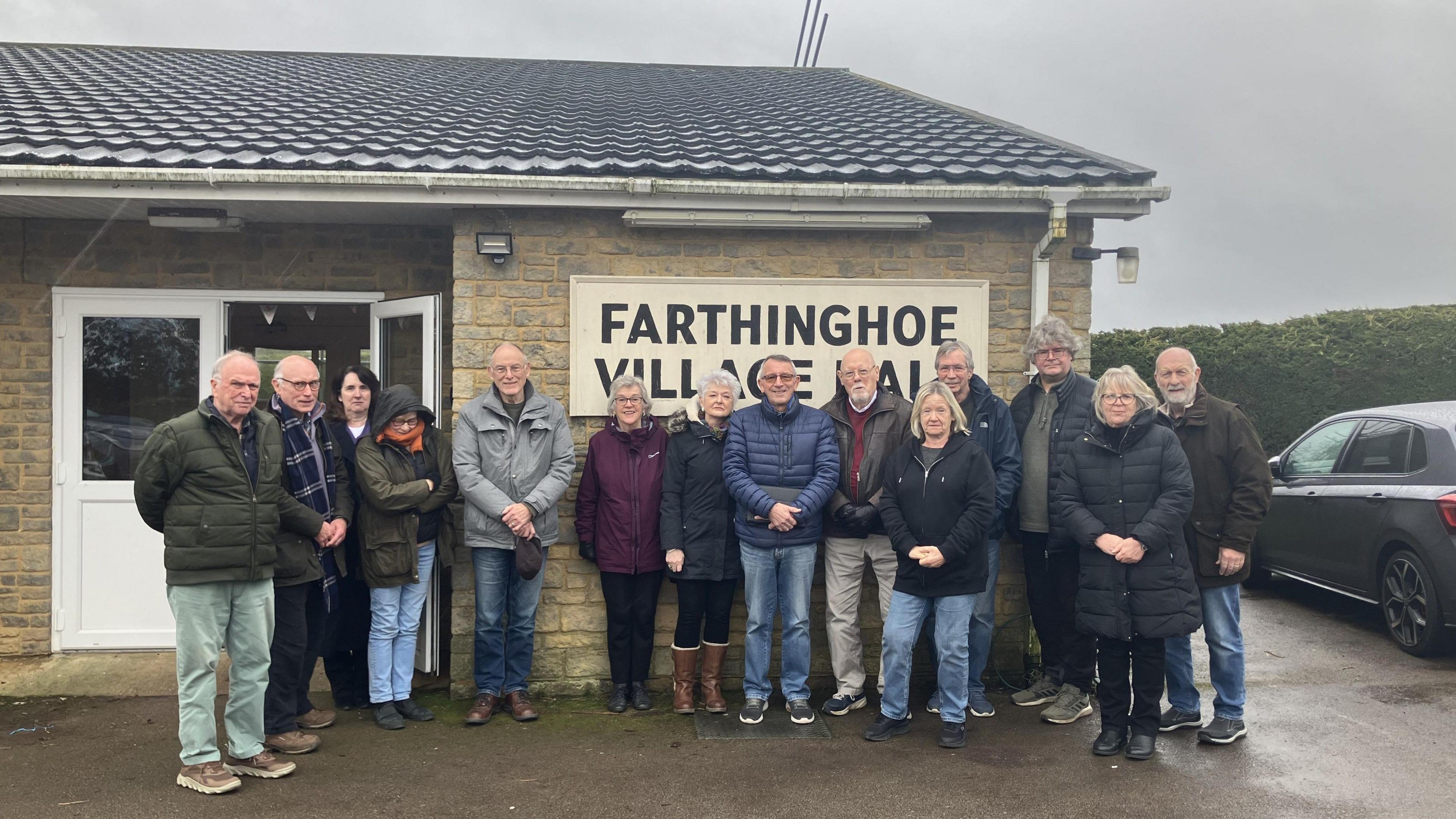 A group of people stand outside a small brick building. 