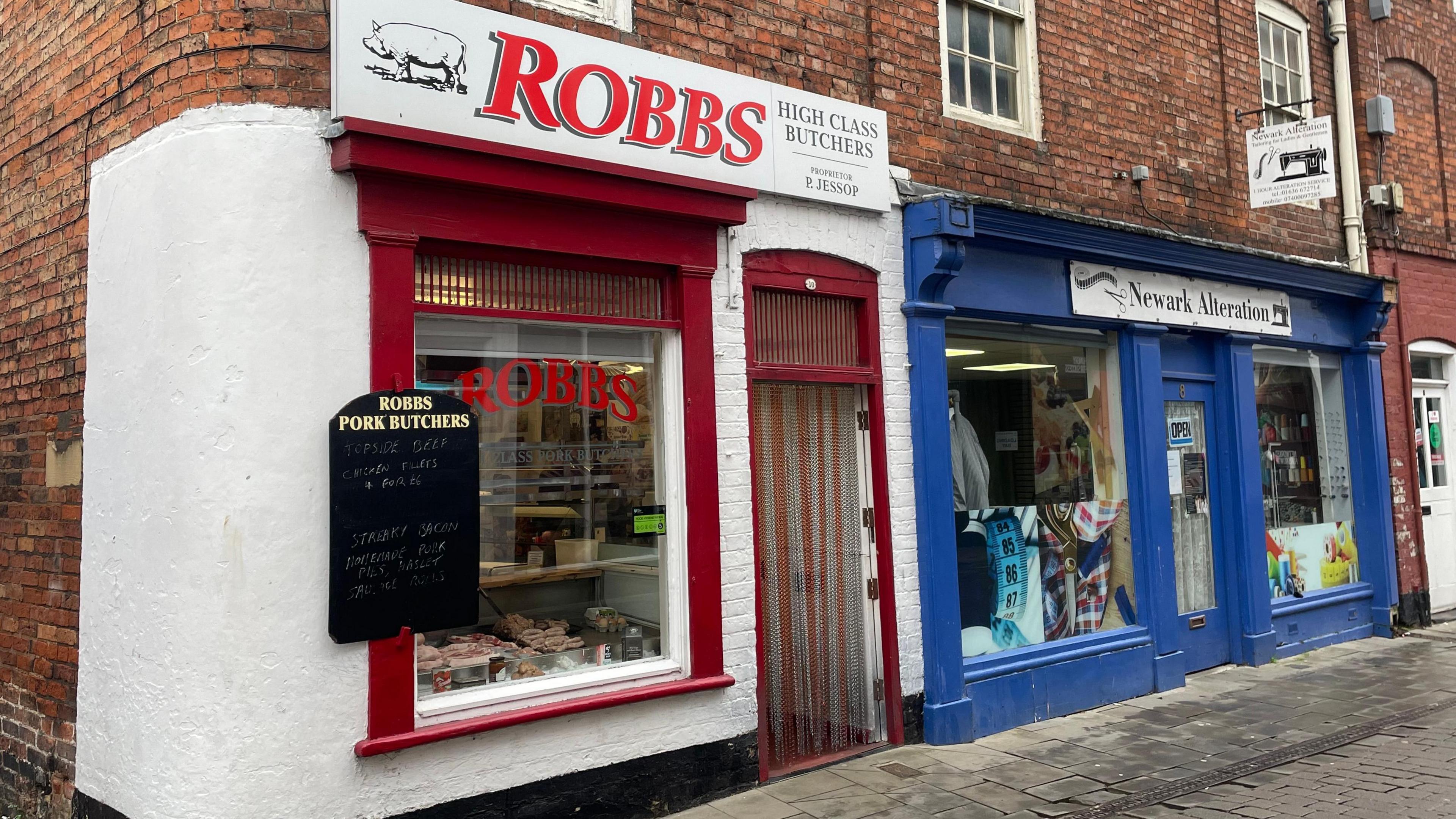 Exterior shot of the white and red painted butcher's shop, consisting of a single large window with a door beside it, which stands on the corner of a row of old-fashioned brick shops.