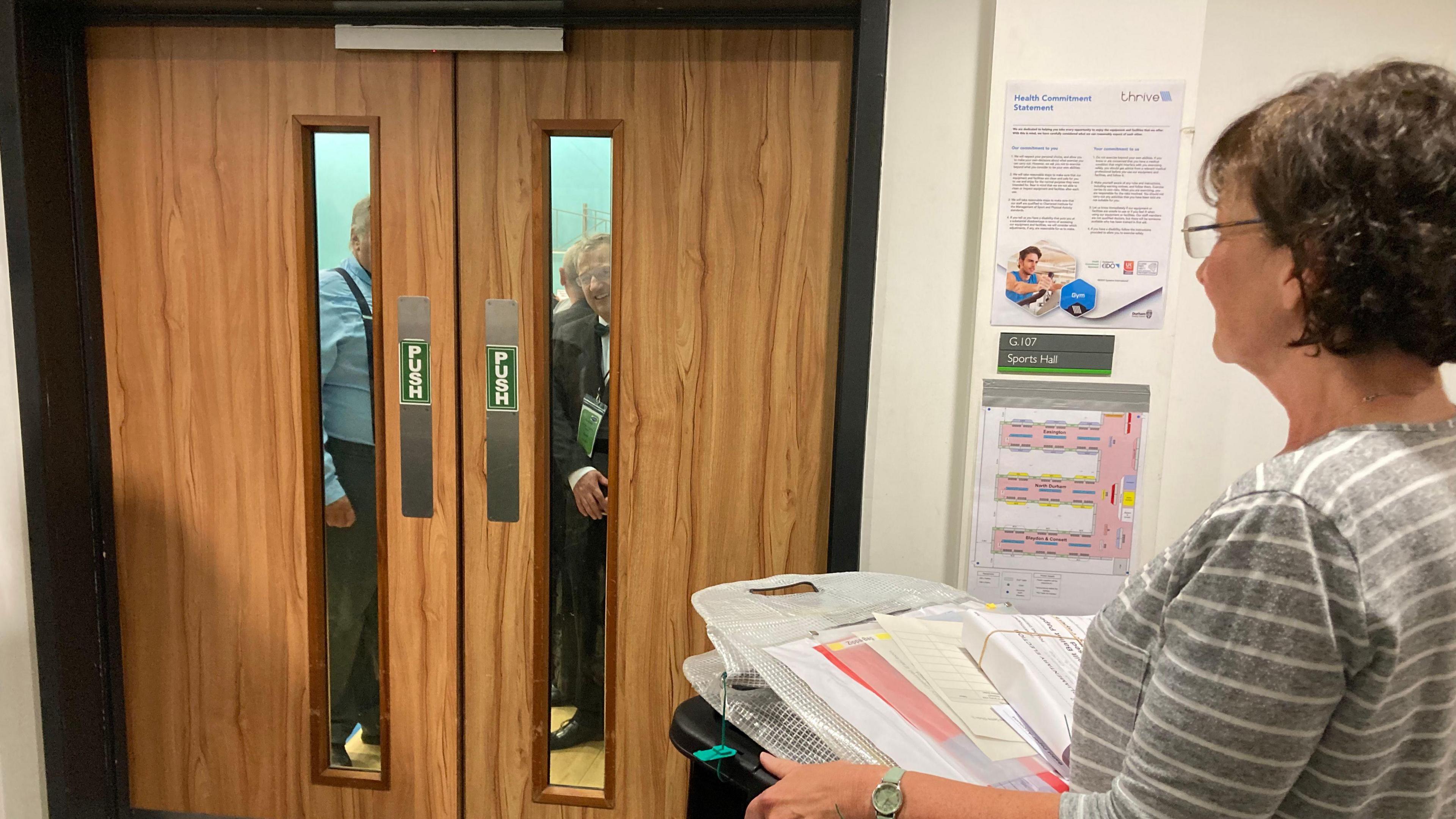 A member of the counting team holding a ballot box in front of a locked door in Consett, with people laughing behind the door