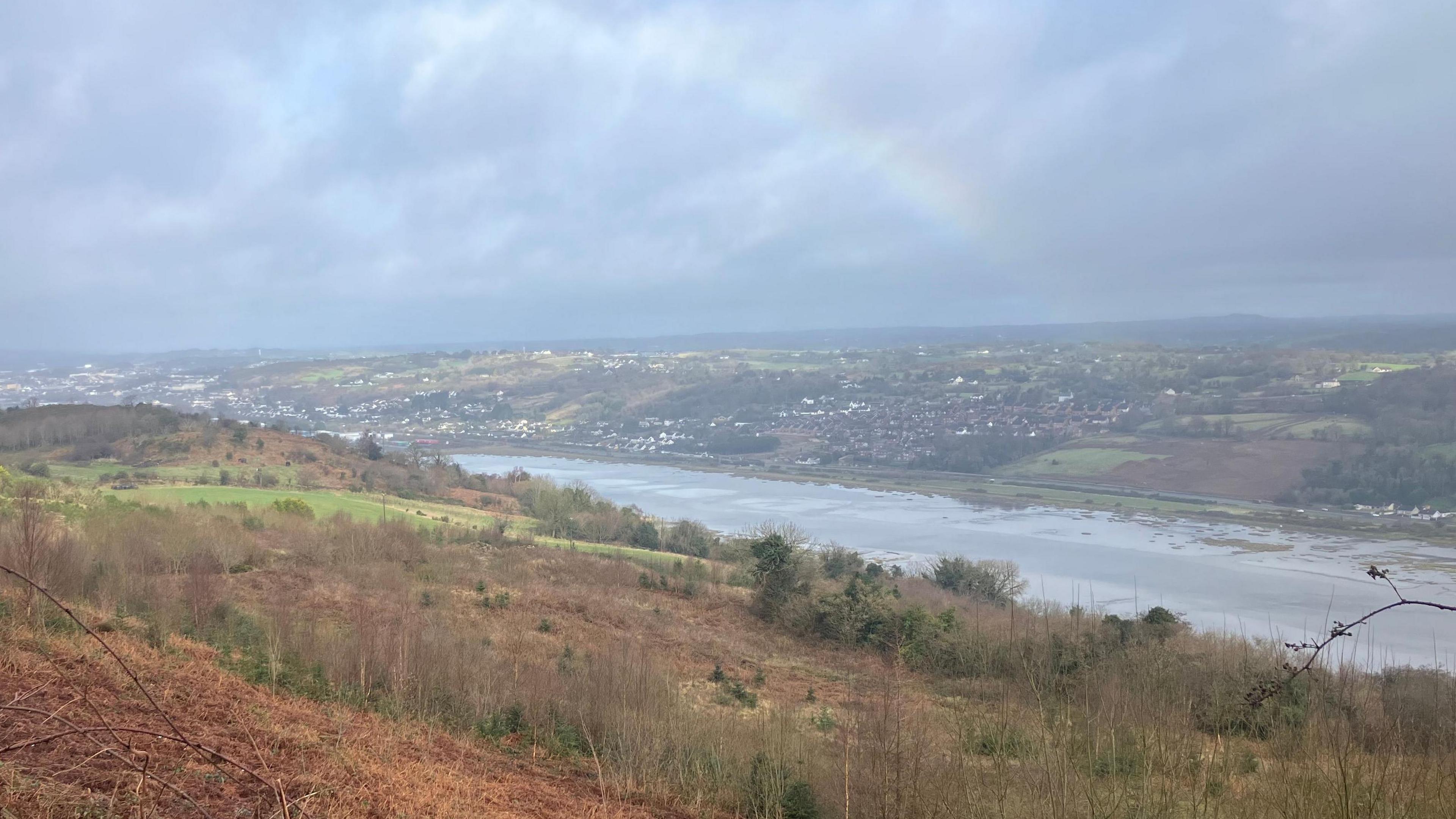 A river divides two pieces of land, one is brown and grey with trees and bushes on it. The other side in the distance is filled with houses and buildings, as well as patches of trees. The sky is a dark grey and there is a faint rainbow over the top of the buildings on the other side of the river.