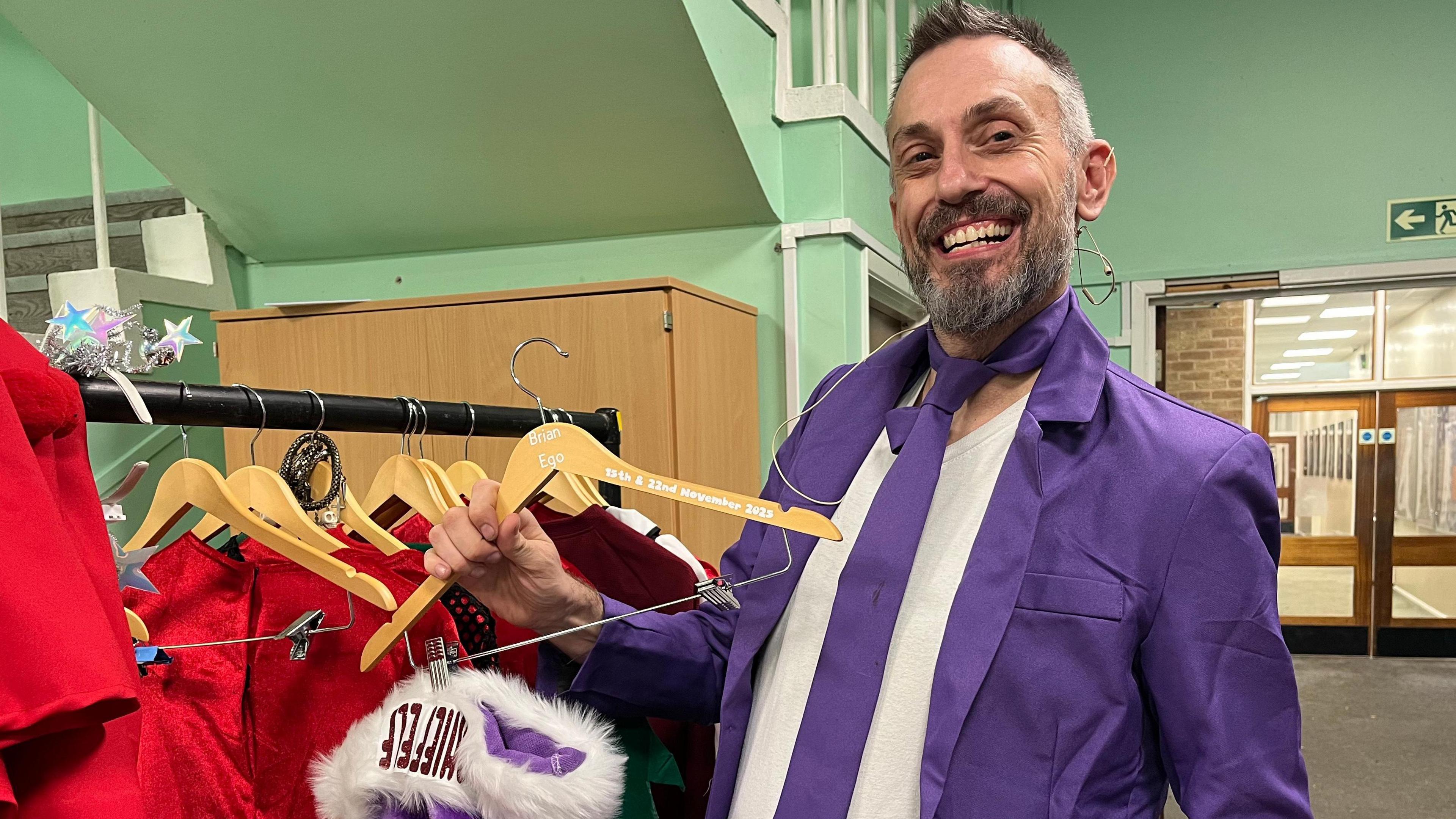 Brian Reeve-Hayes is wearing a purple suit and holding a purple Christmas hat. He is smiling at the camera. He has greying hair and beard.