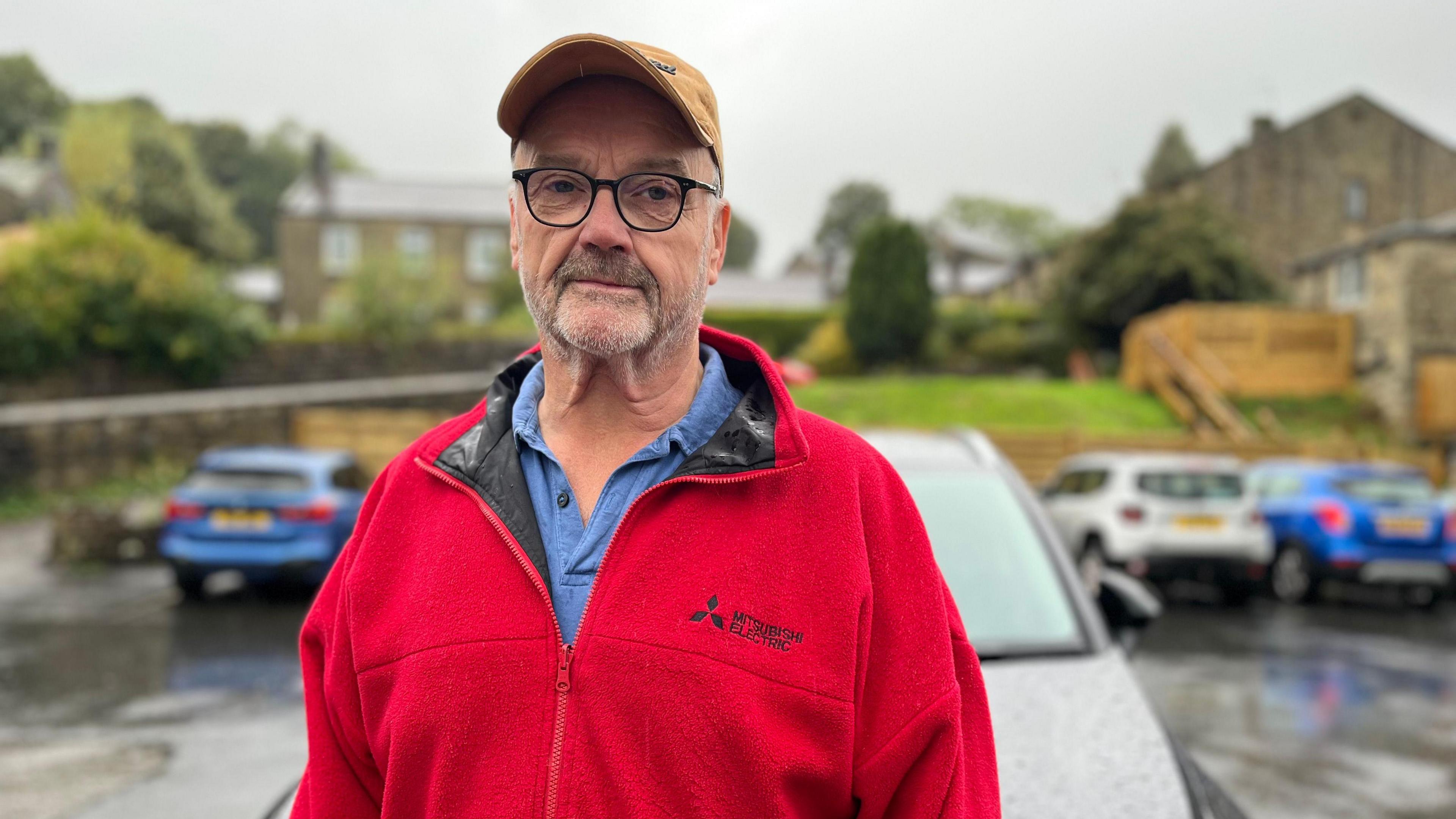 A man wearing a cap, and red fleece over a blue shirt, stands in front of a black car in a petrol station car park.