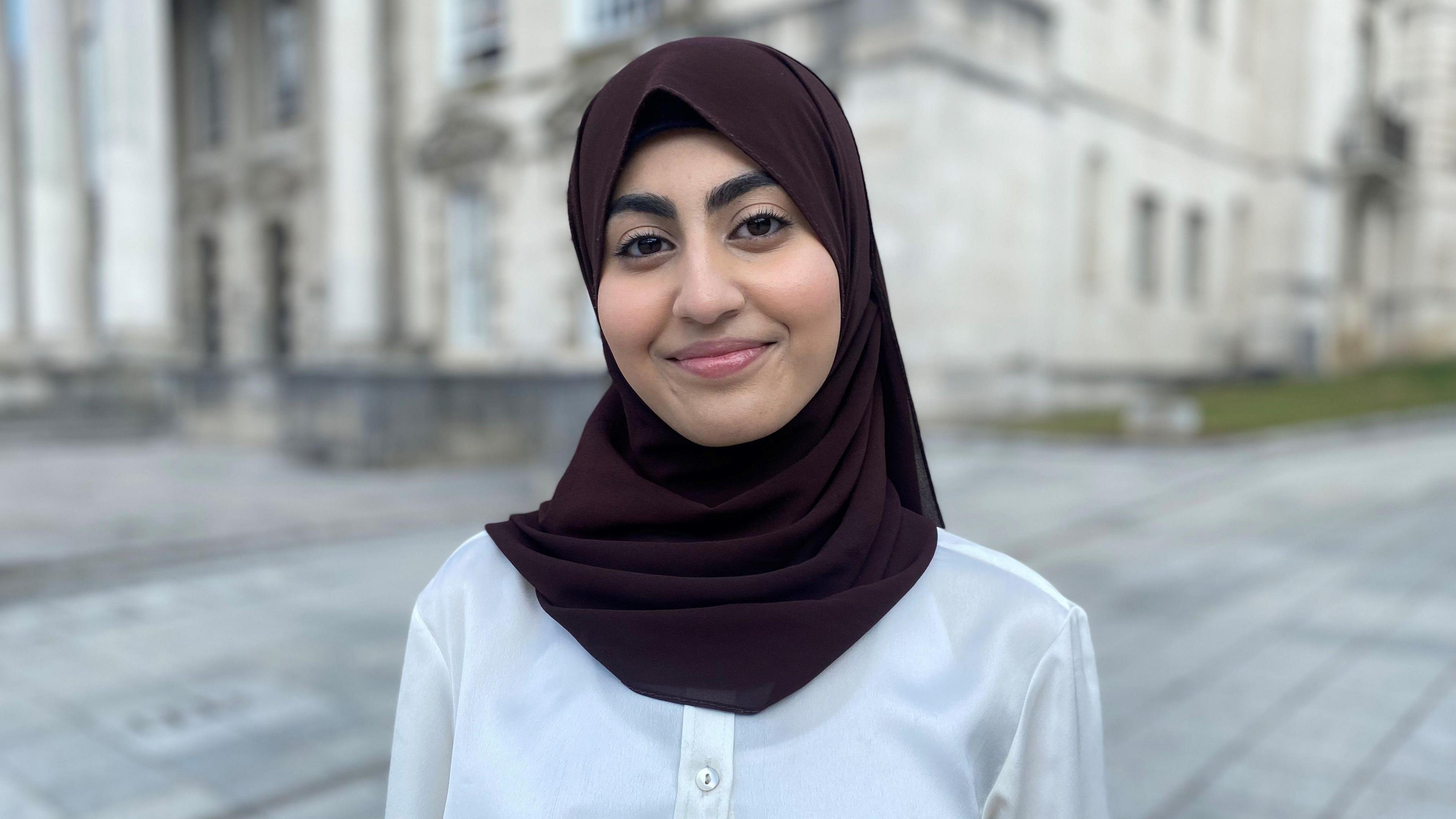 A teenage girl wearing a head covering smiles at the camera. She is stood near a white civic building in Leeds.