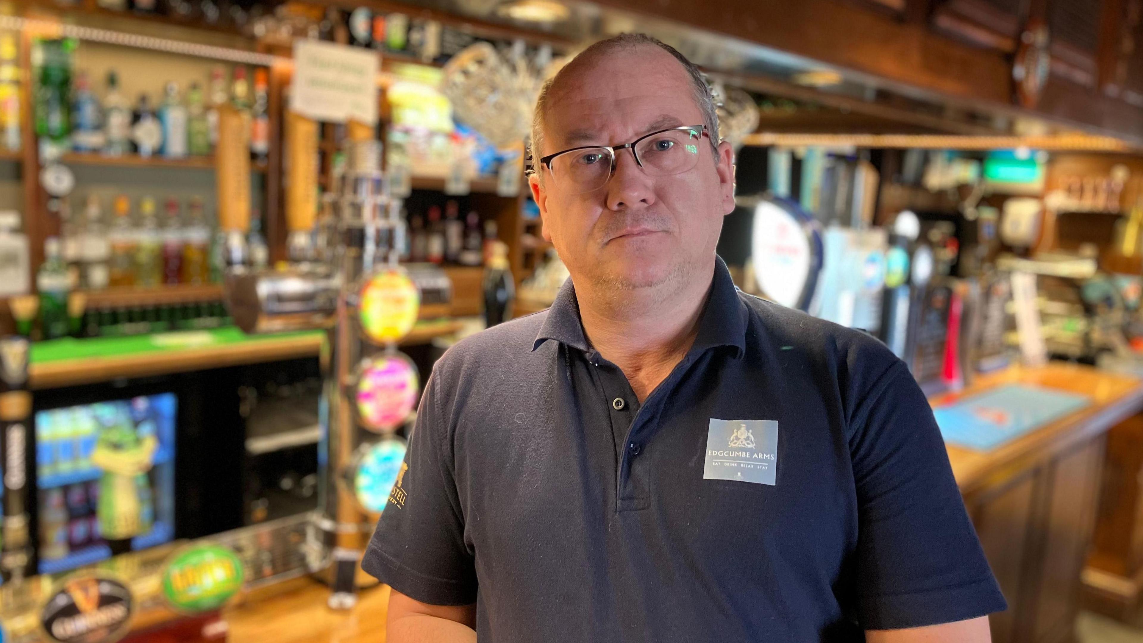 A man in a black t-shirt stands in front of a brightly lit bar in a pub.
