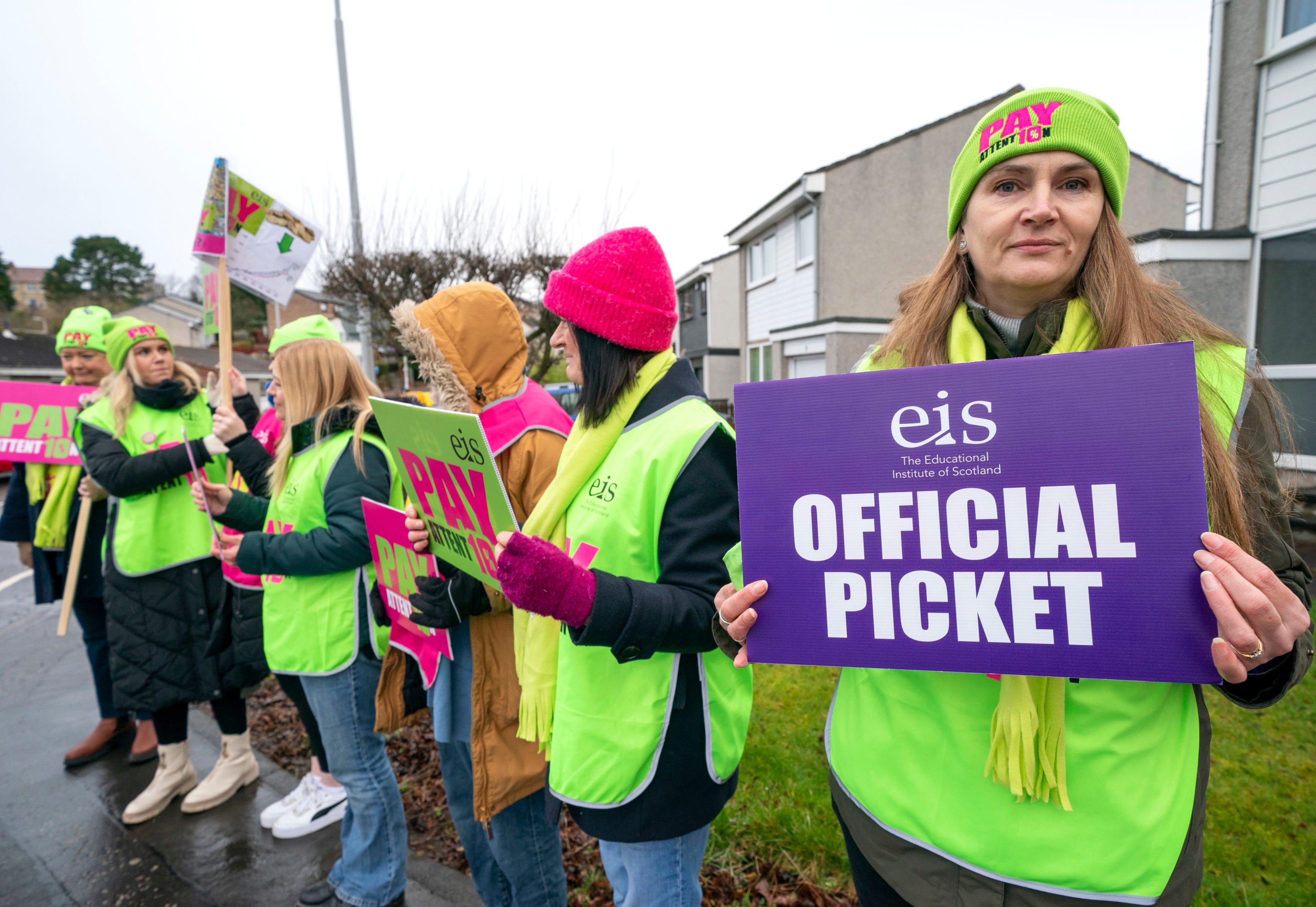 A group of  seven teachers stand in a line on an EIS picket line in a street in Dunfermline. The closest to camera on the right of the image is holding up an "EIS official picket" placard.