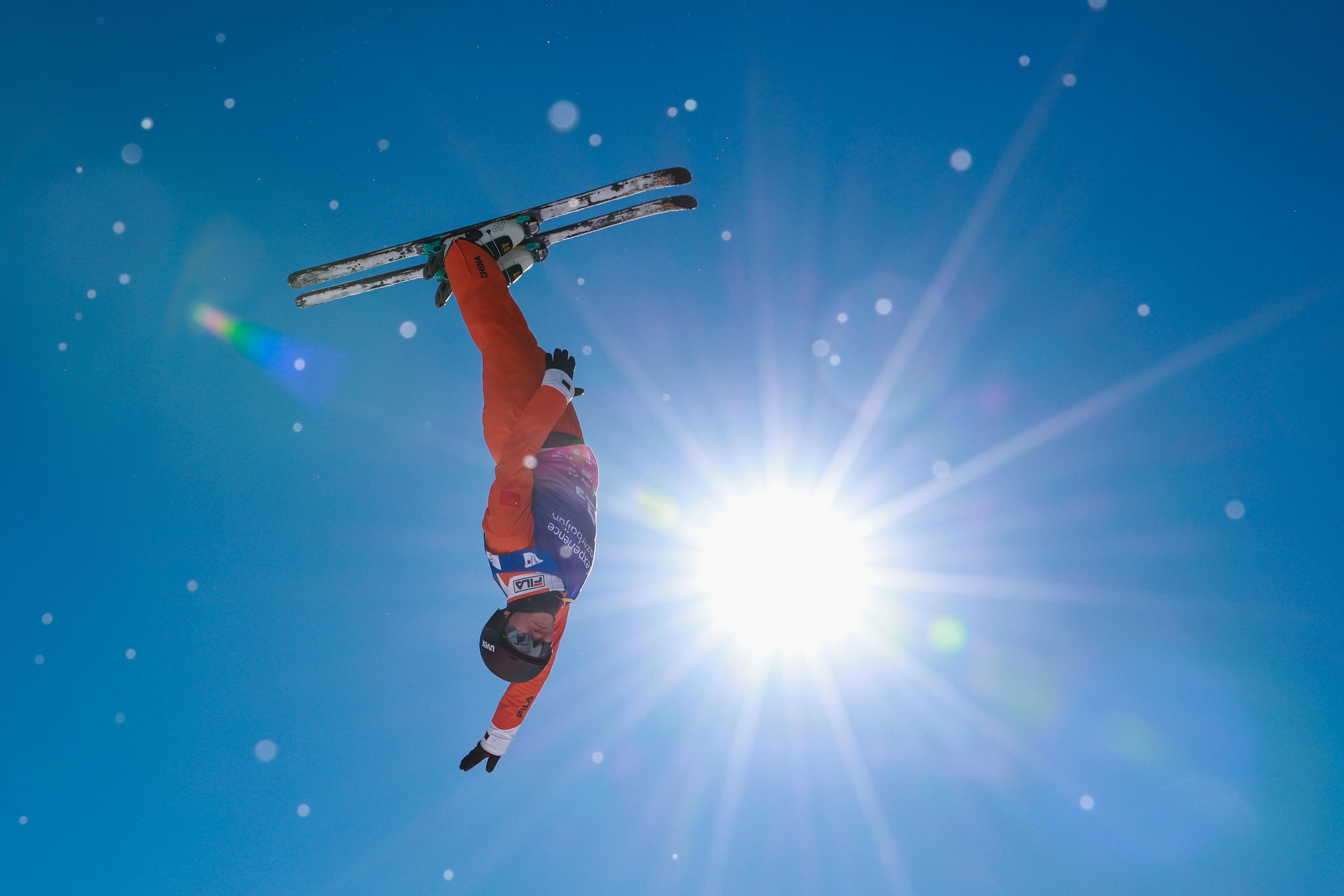 A skier in a bright orange suit performs a dramatic inverted aerial maneuver against a clear blue sky, with skis pointed upward and one arm extended toward the ground. The sun shines intensely behind the athlete, creating a burst of light and lens flares across the image.