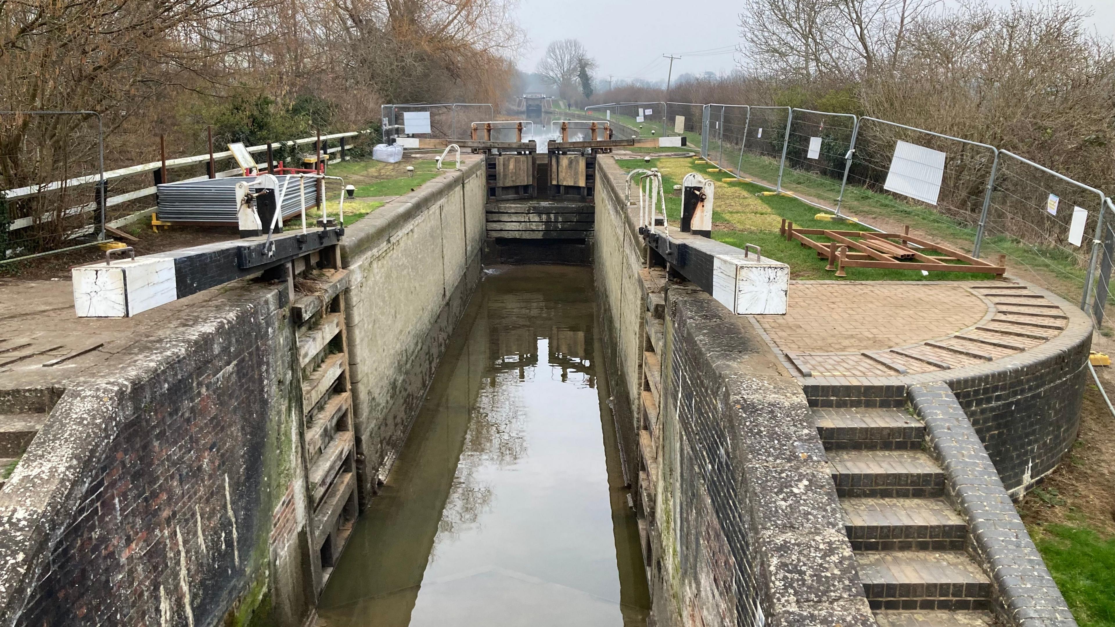 A lock on a canal. The gates nearest the camera are open, whilst the others are closed with a board beyond to prevent usage. There is a shallow water pool remaining in the lock, and the shadow of the lock gates is visible in the water. There is a metal fence to prevent access to the site. Stone steps from the towpath to the locks are visible to the right.