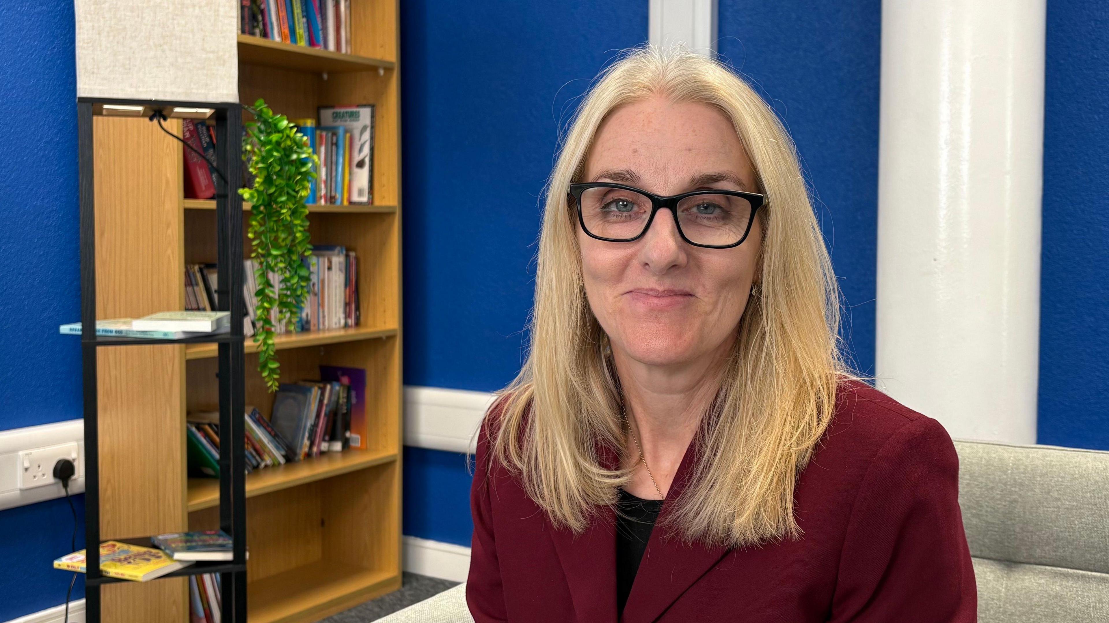 Dr Joanne Summers looking direct to camera. She is sitting inside a room with deep blue walls and a white pillar over her shoulder to the right. On the left behind her is a bookshelf partially-filled with books, as well as a green plant. Dr Summers is smiling and is wearing a dark red jacket over a black top. She has black rimmed, rectangular glasses on and her blonde hair is parted in the middle and is falling over her shoulders.