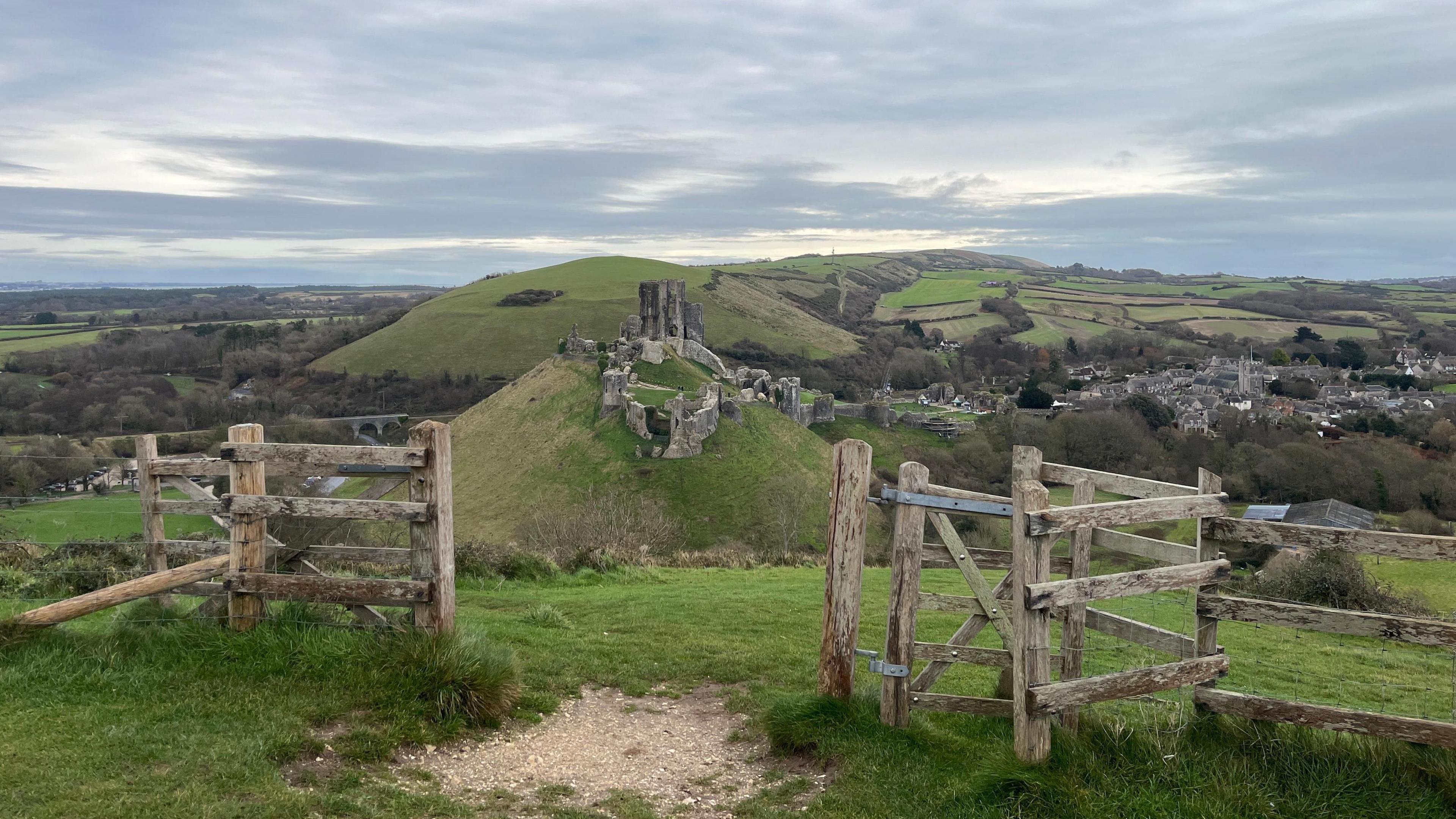 The ruins of Corfe Castle sit on top of a hill in the centre of the picture. In the foreground stands a wooden gate and in the background you can see a patchwork of fields stretching to the horizon.