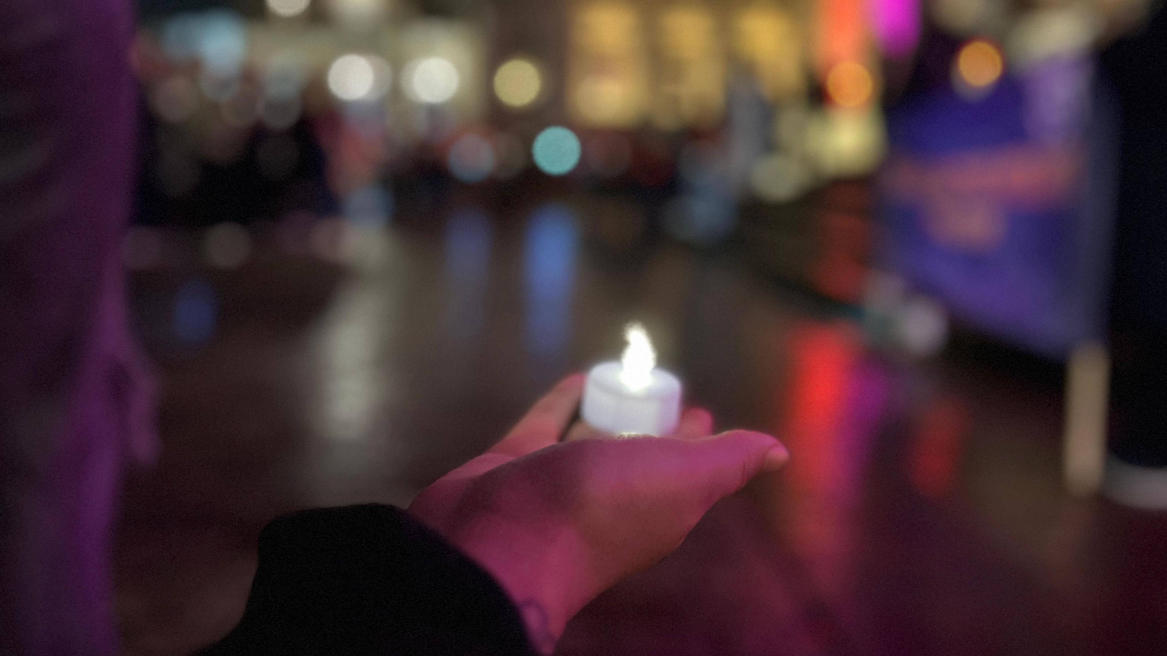 An electronic candle is held on the palm of a hand, the background is blurred.