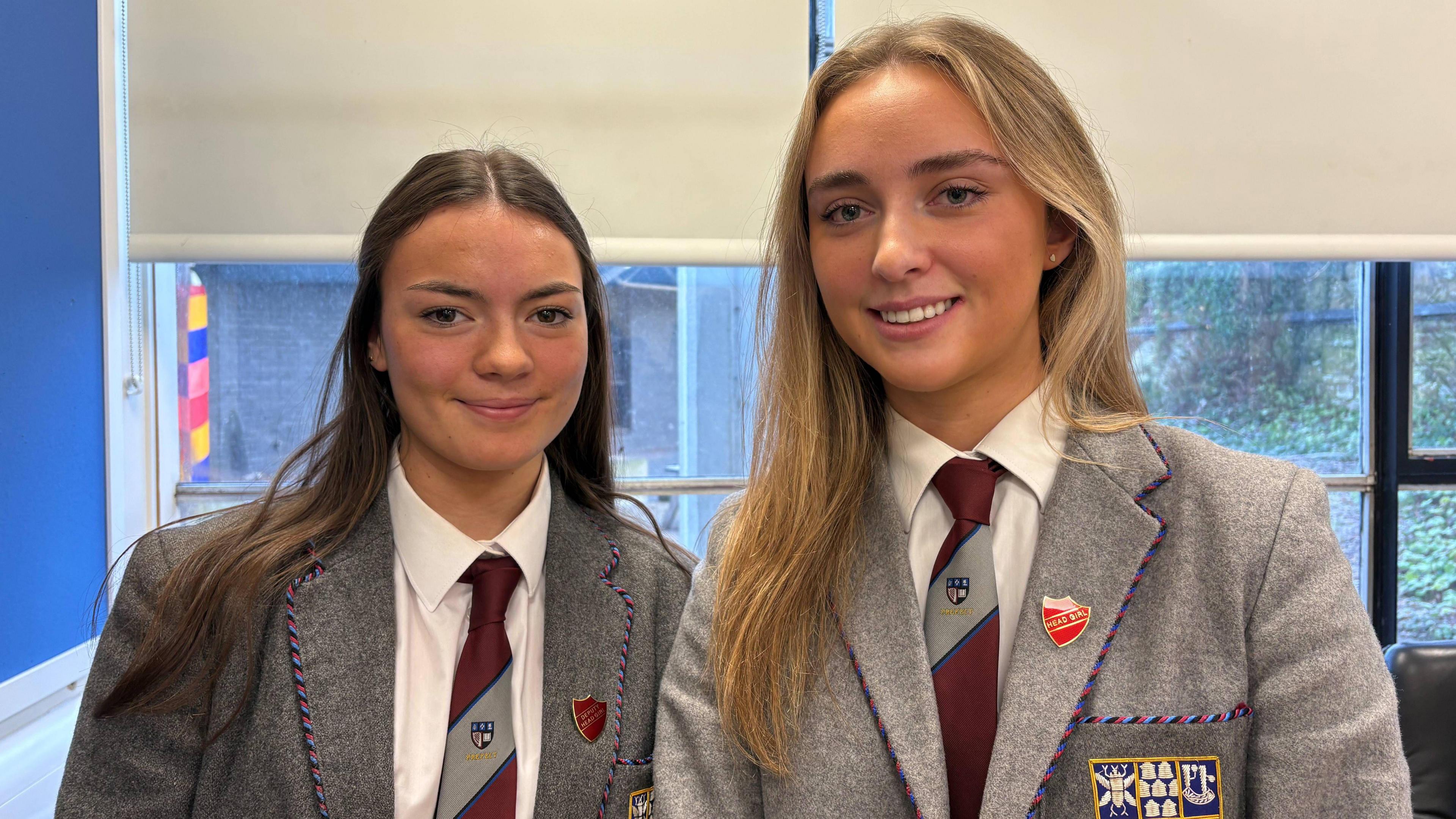 Charlotte and Olivia smile in their grey school uniform. They both wear badges and have a maroon and grey tie with a white shirt.