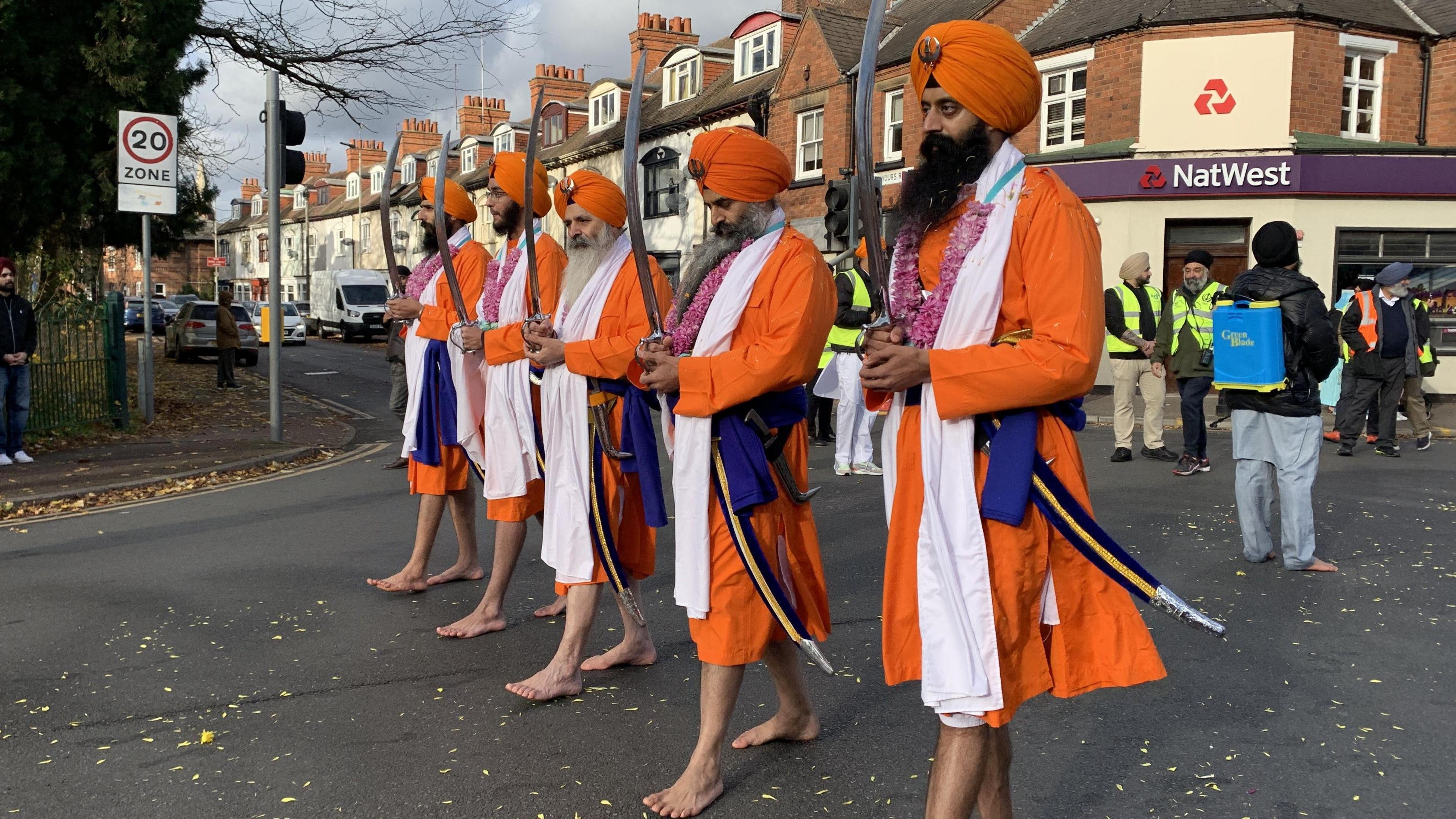 Five men in traditional orange dress holding swords vertically and walking side by side bare foot