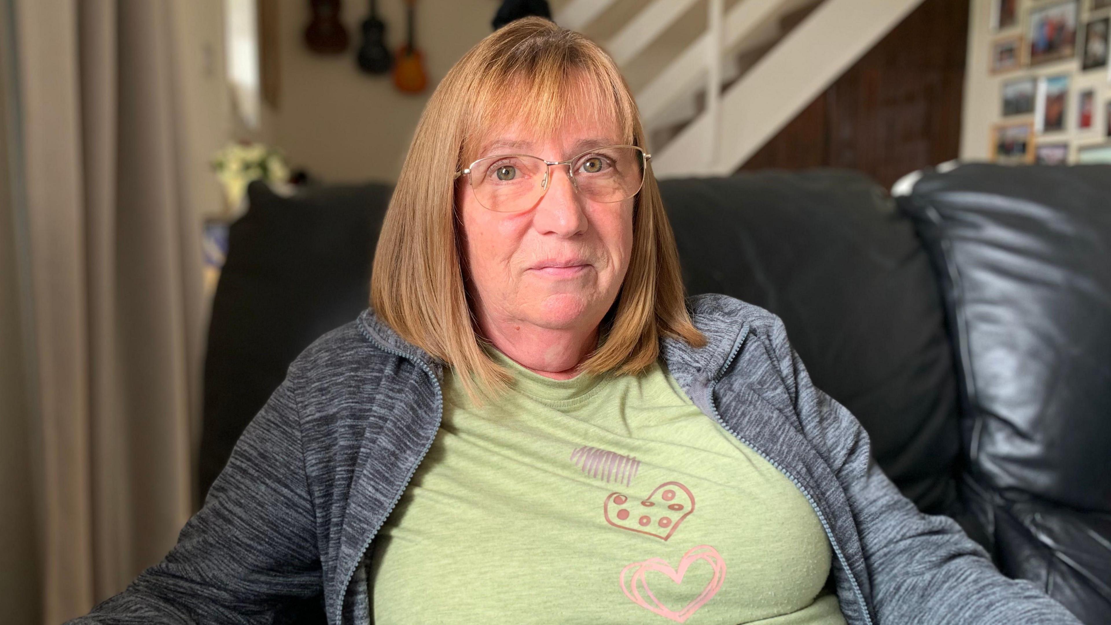 A woman with brownish-red medium length hair in glasses in a grey cardigan with a green T-shirt with heart logos on it. She is sitting on a leather armchair with a staircase going up behind her.