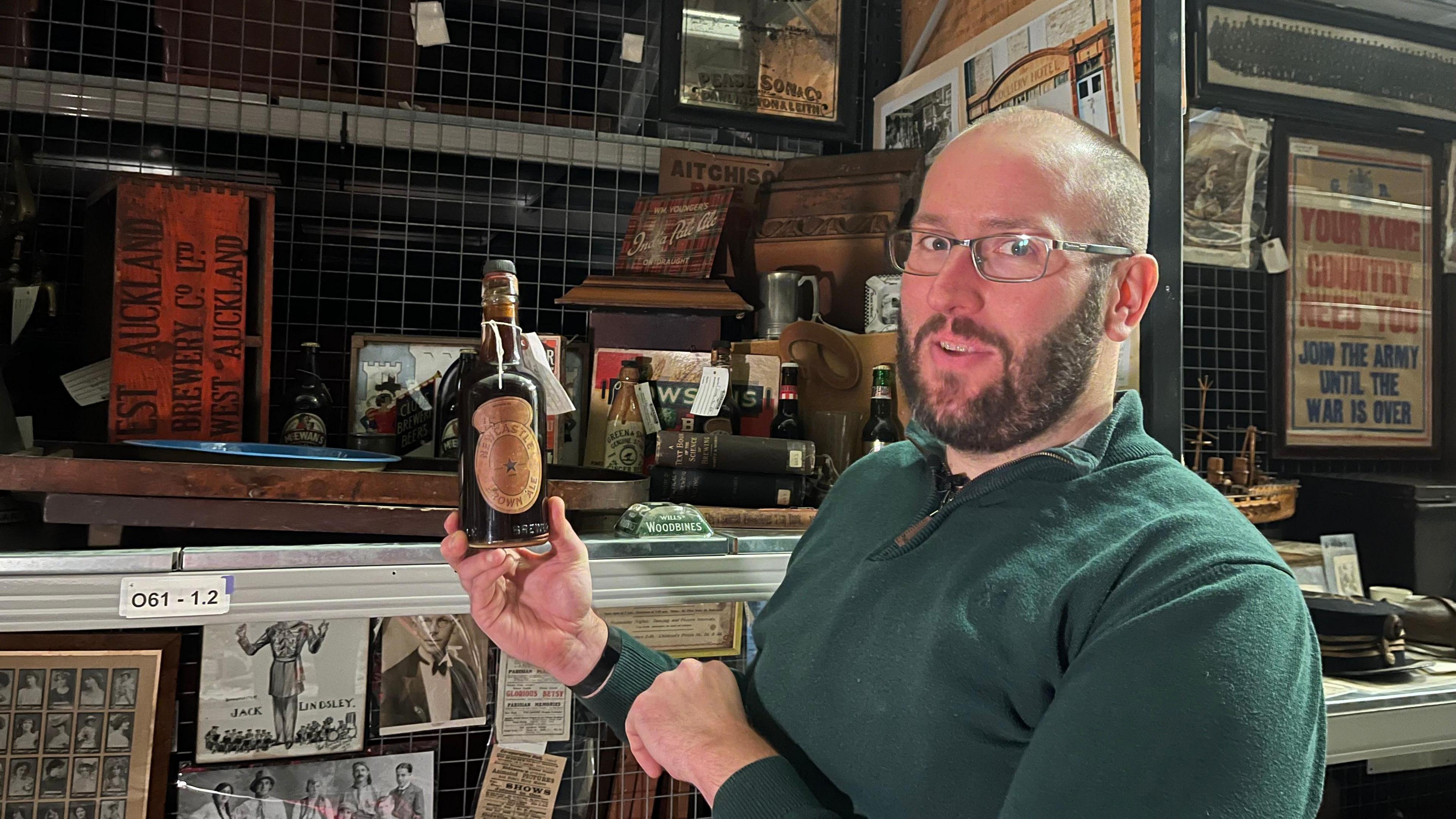 Dan Hudachek wearing a green jumper and glasses. He is holding a 1928 bottle of Newcastle Brown Ale. In the background are shelves filled with objects and photographs. 