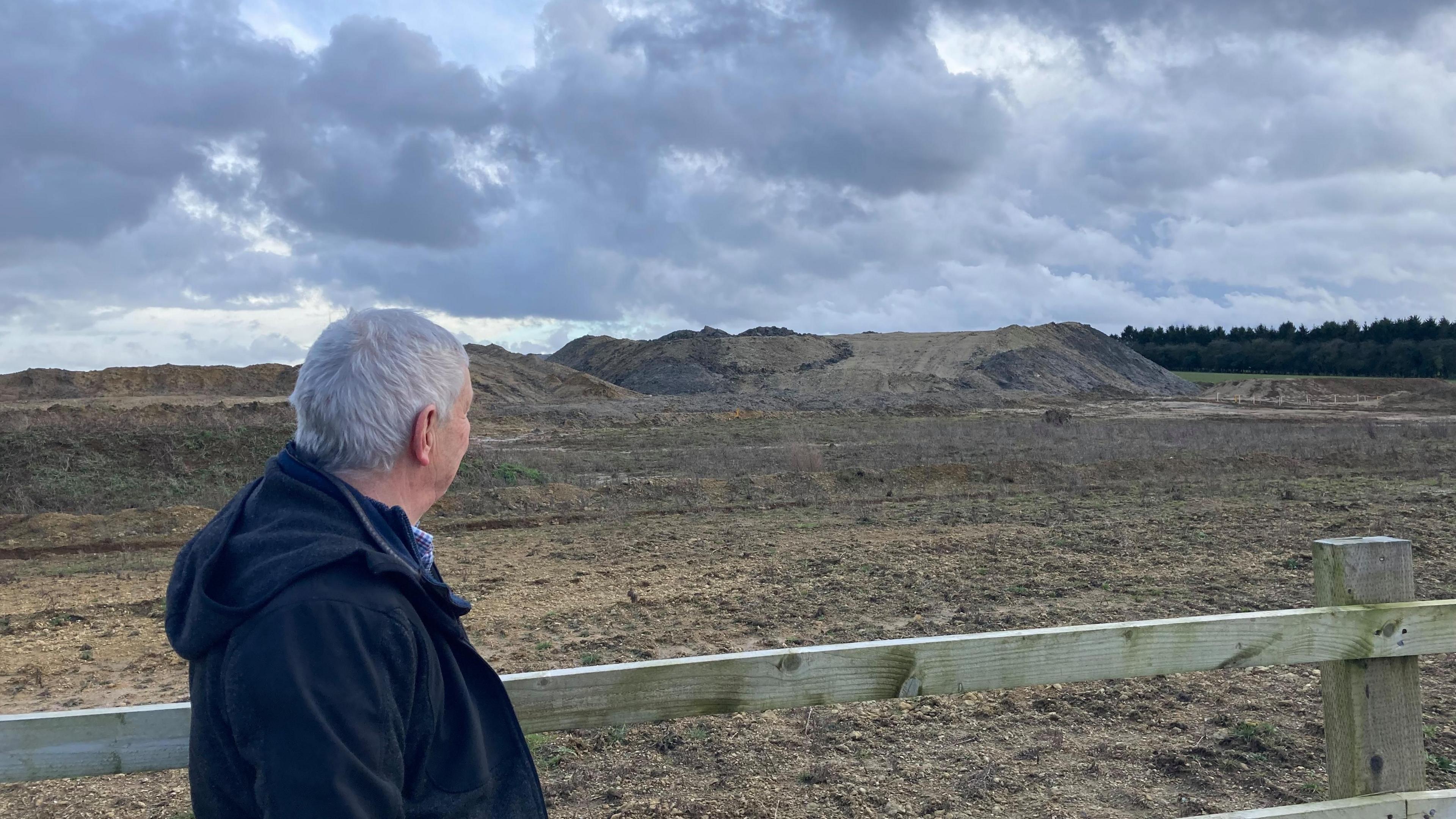 Stephen Adkins standing next to a fence looking over his shoulder at a mound of mud in the distance