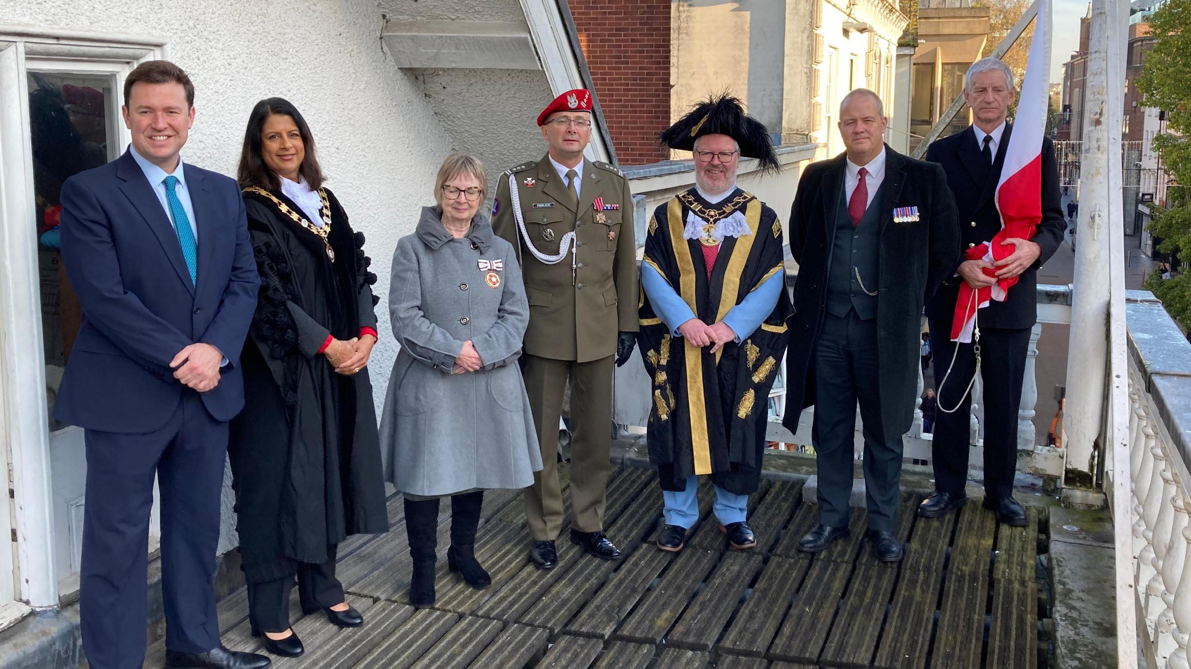 L-R: Exeter MP Steve Race, Exeter City Council chief executive Bindu Arjoon, Deputy Lieutenant for Devon Peta Foxall, Robert Pawlicki - defence attaché for the Polish Embassy, Exeter's Lord Mayor Councillor Kevin Mitchell, Dr Simon Selby - honorary consulate for the Republic of Poland and John Davies - mace sergeant at Exeter City Council. They are all standing on a balcony at Exeter's Guildhall during a Polish flag raising ceremony on a sunny day. 