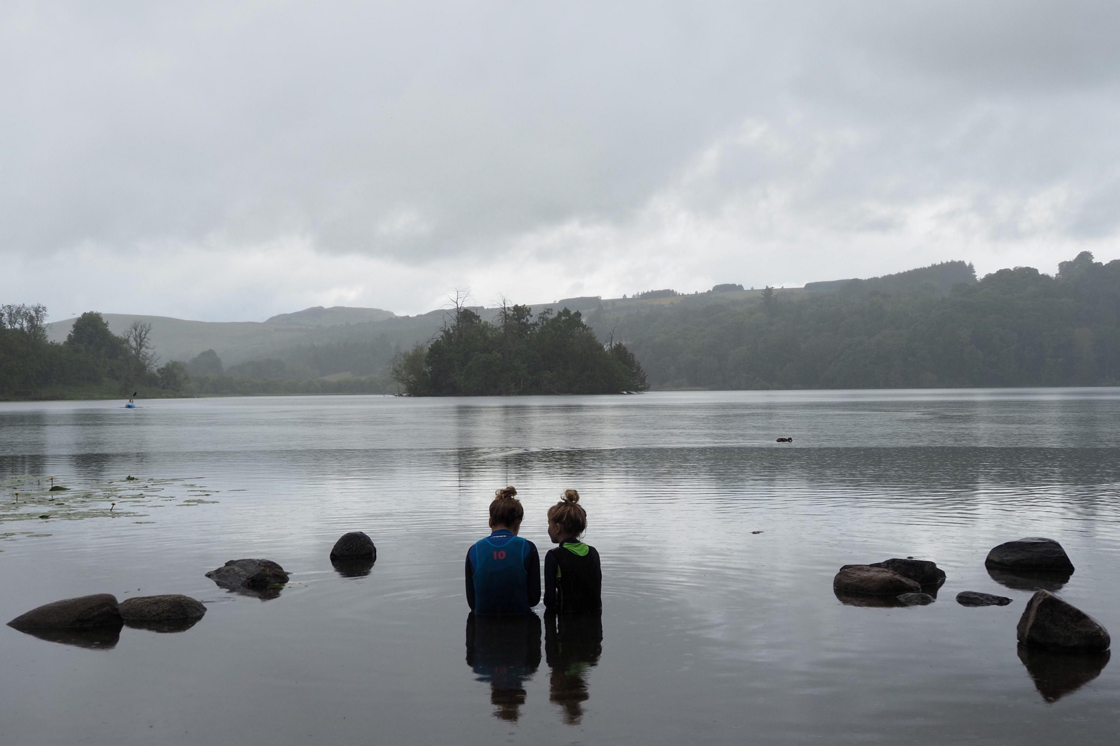 Two young girls sitting next to each other at the edge of a loch.