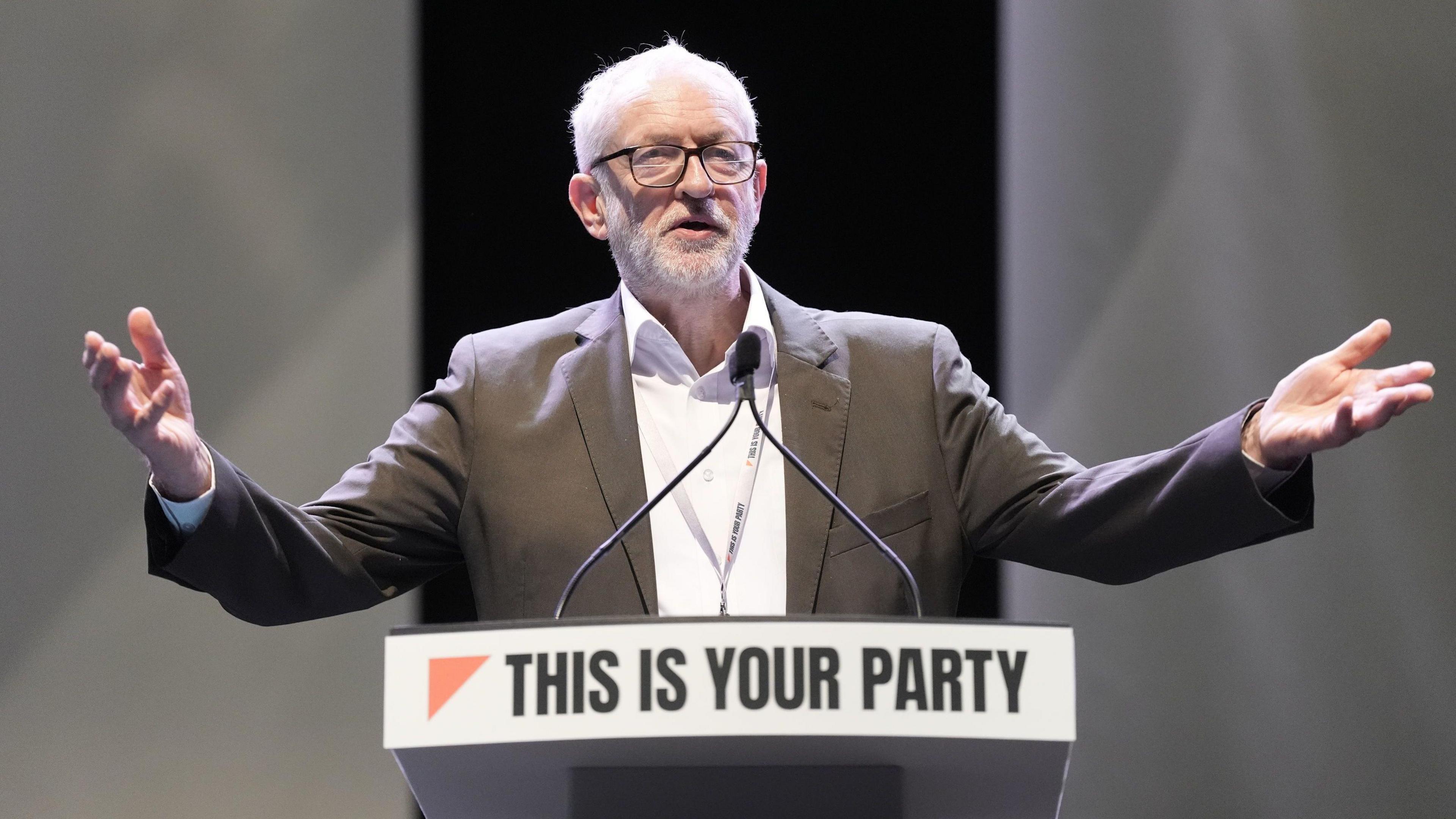 Jeremy Corbyn stands behind a podium on a stage with arms outstretched. The podium has a sign reading ‘THIS IS YOUR PARTY’ in bold black letters with a red triangular graphic on the left. Two microphones are mounted on the podium, and the background is dark with soft lighting.