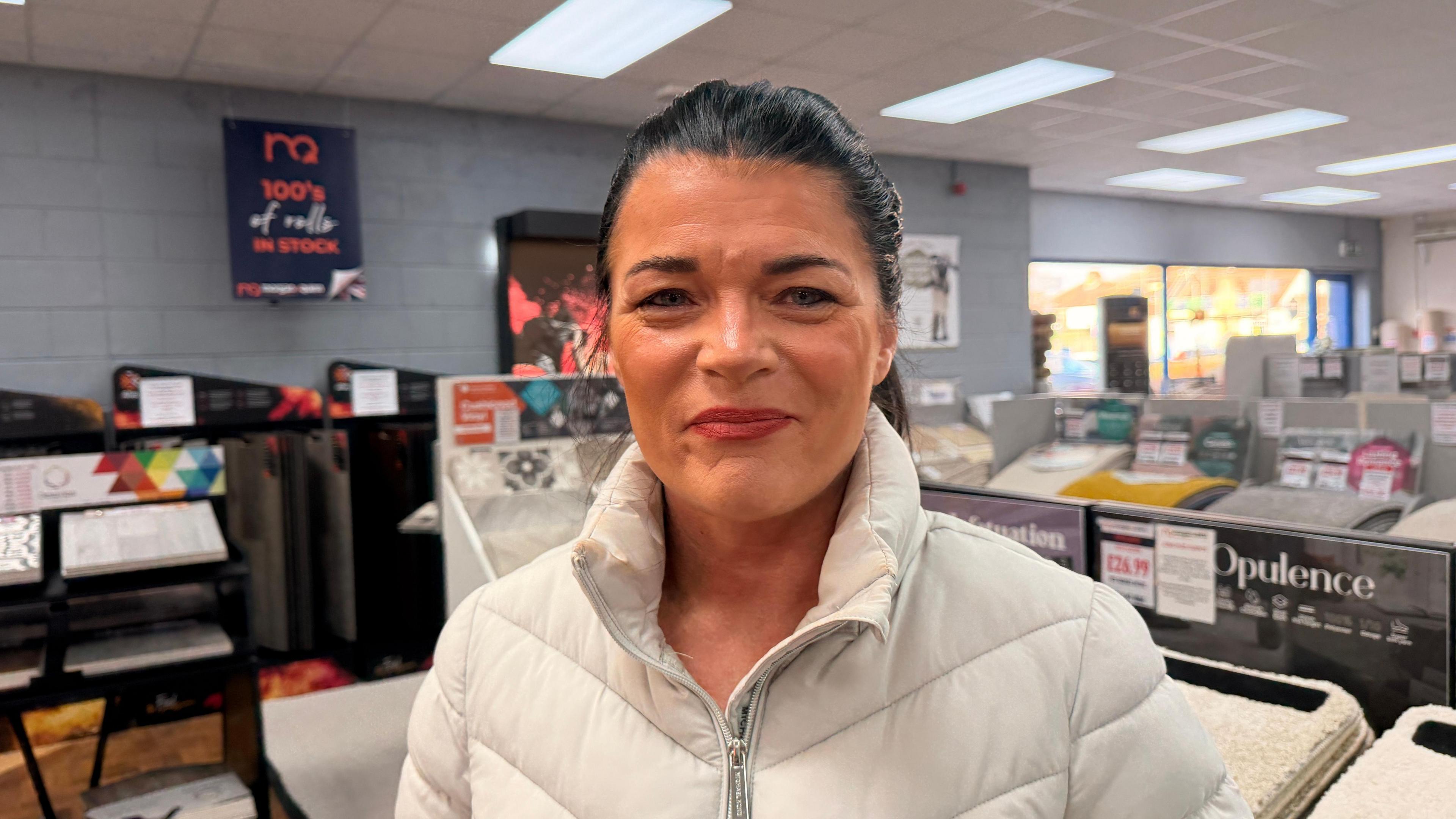 A lady in a white jacket in a carpet shop, with black hair and makeup on. She is the manager at the store 