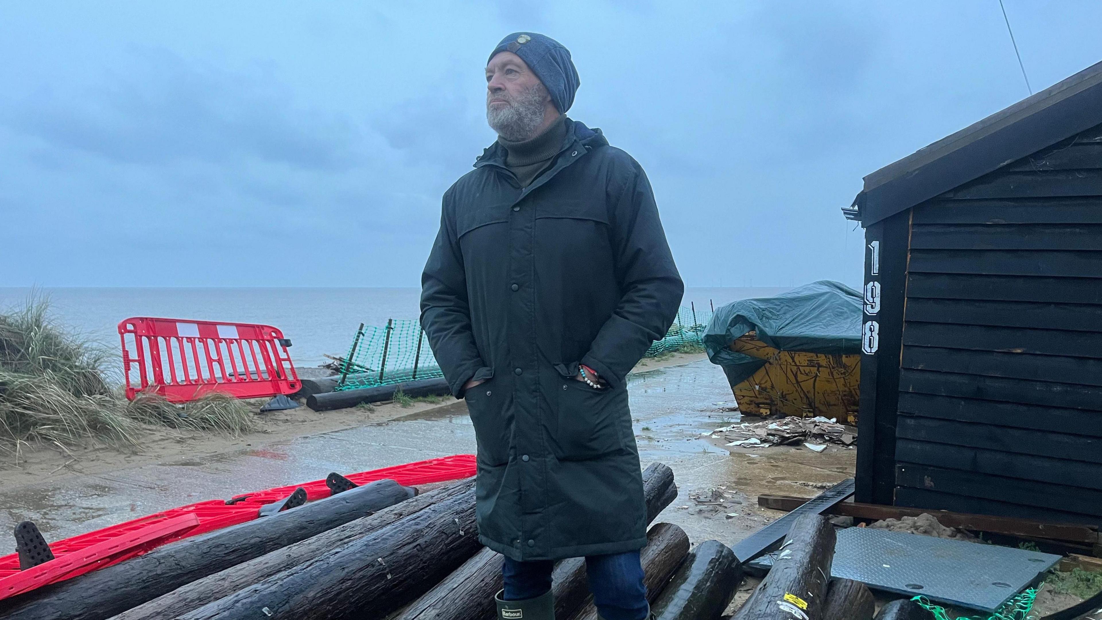 Lance Martin wearing a dark green, knee-length waterproof coat and a blue beanie hat, with a close-cropped grey beard, looking away, with his hands in his coat pockets, standing beside a pile of old telegraph poles and behind him the sea to his left and a skip and a black timber-clad house on his right