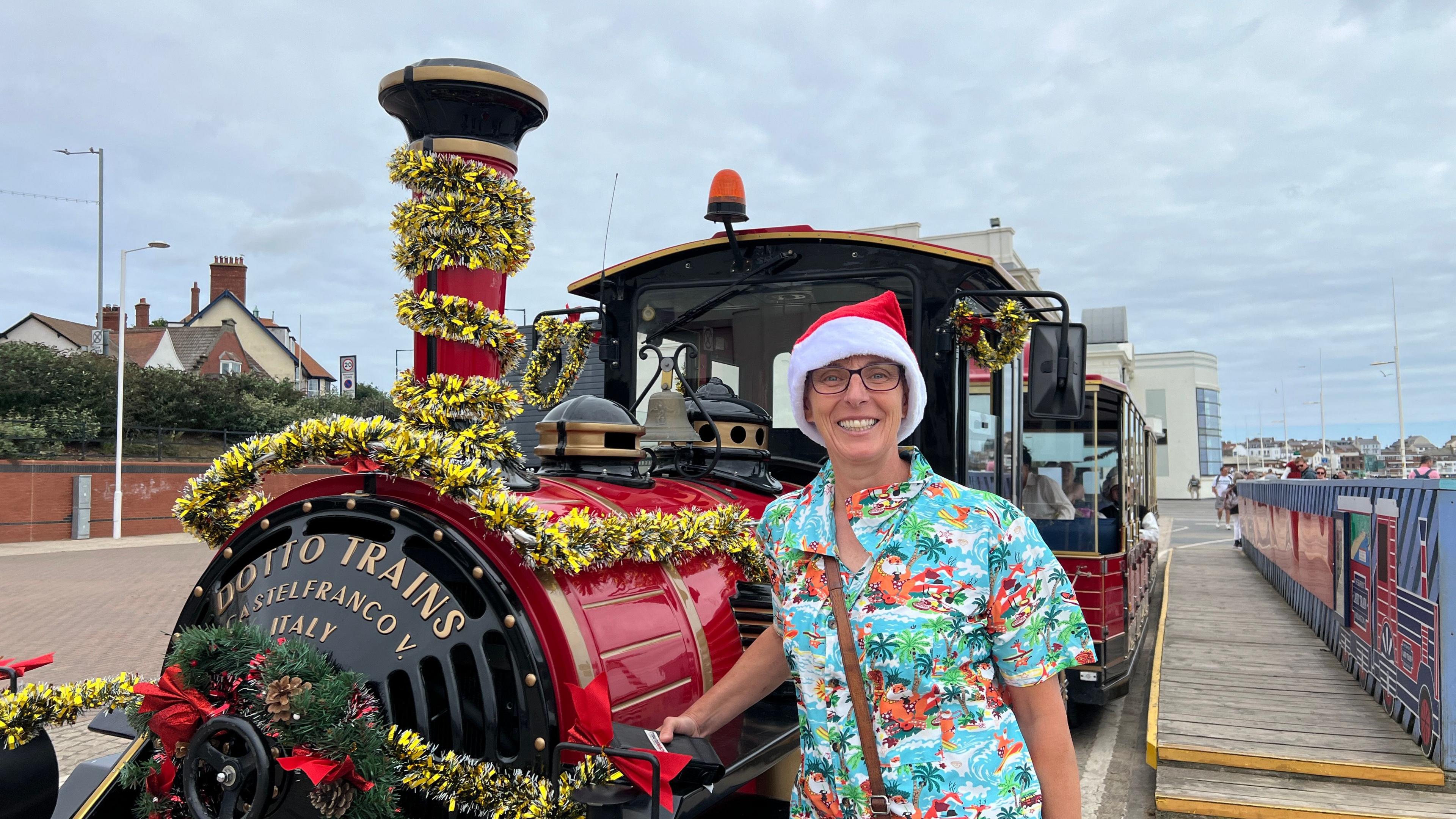 A woman wearing a Hawaiian shirt and a Santa hat stands next to a land train covered in Christmas decorations