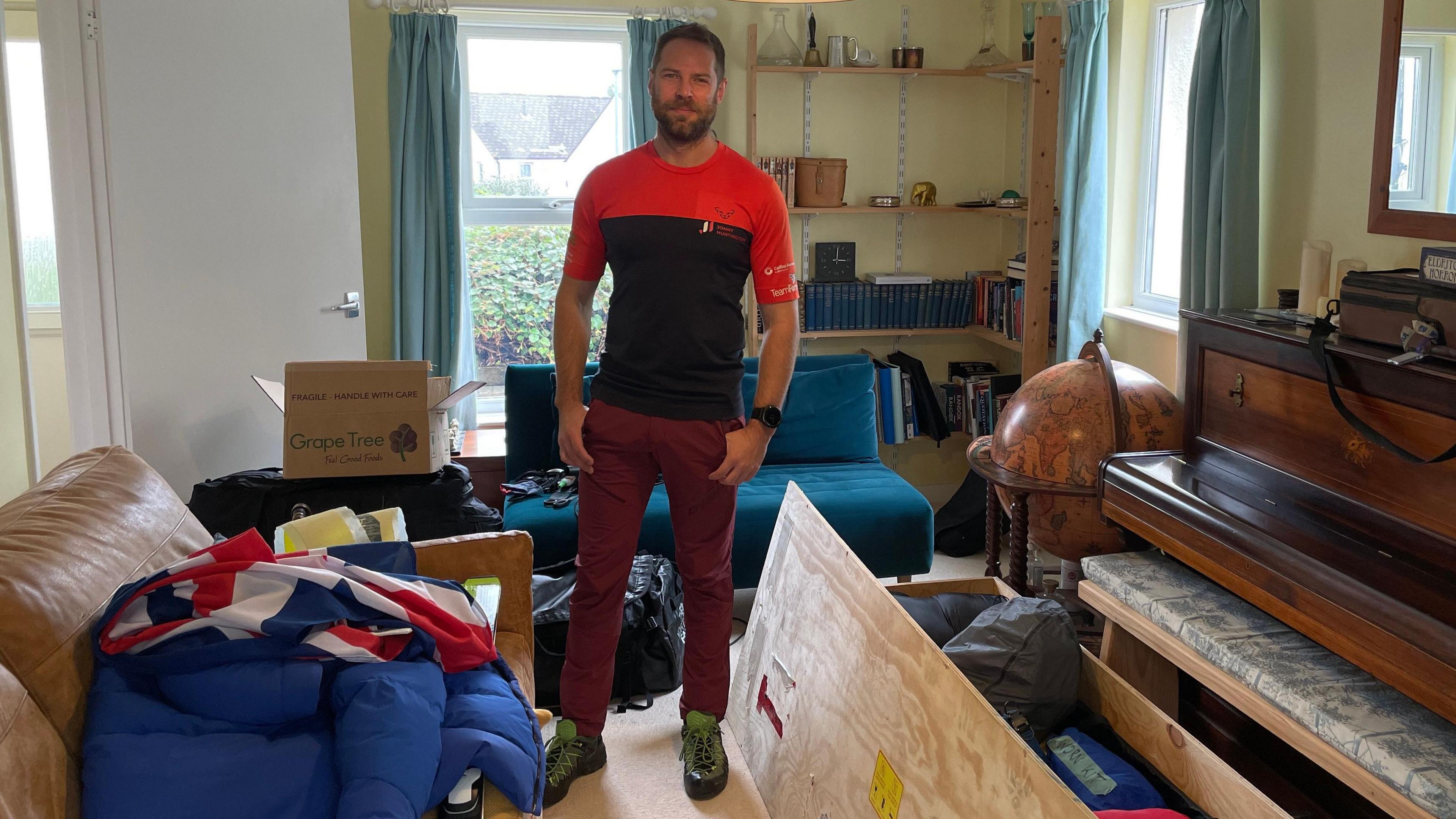 Jonny, standing in a living room, is wearing a half-red, half-black T-shirt and burgundy trousers. There is a sofa, a piano, a blue futon and a globe. He is surrounded by bags on the floor which contain his kit.