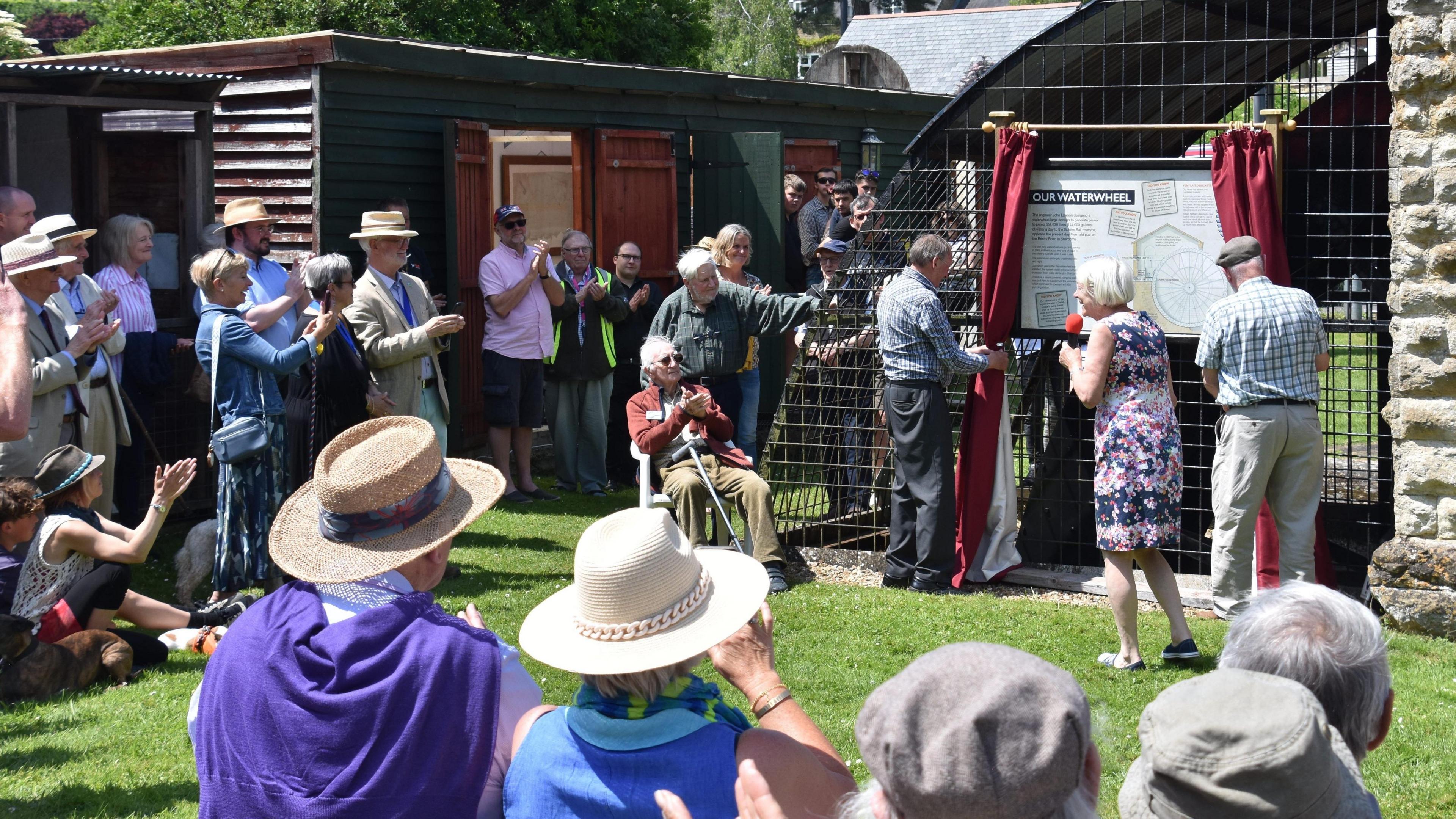 A group of people standing around a half submerged waterwheel enclosed in a cage. On the cage is a pair of red curtains framing an information board. It's a sunny day and some of the people are wearing brimmed hats.