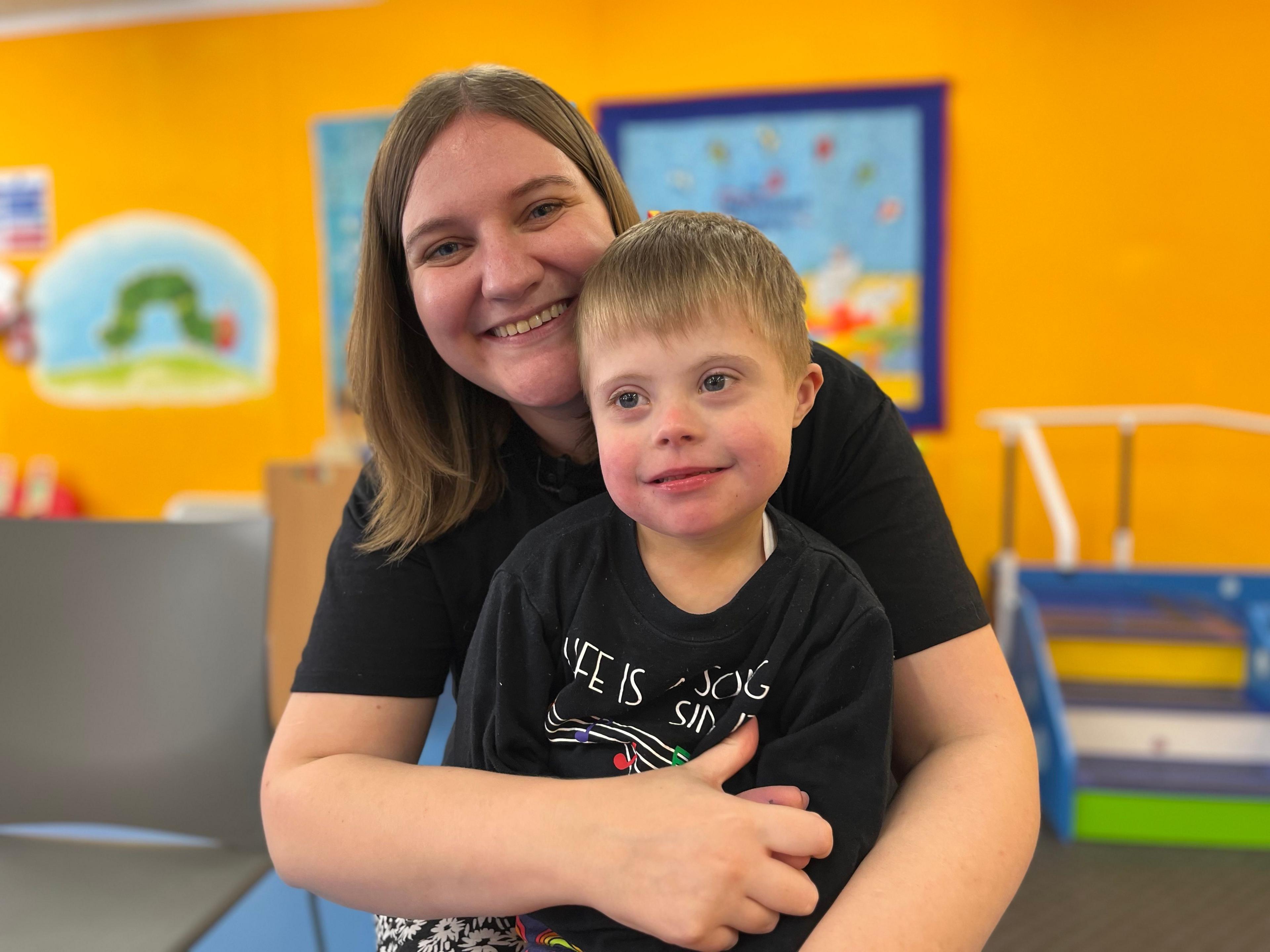 Jaxson is sat on his mum Clare's lap, she has her arms wrapped around him and they are both smiling. They are in a brightly coloured room in The Movement Centre.