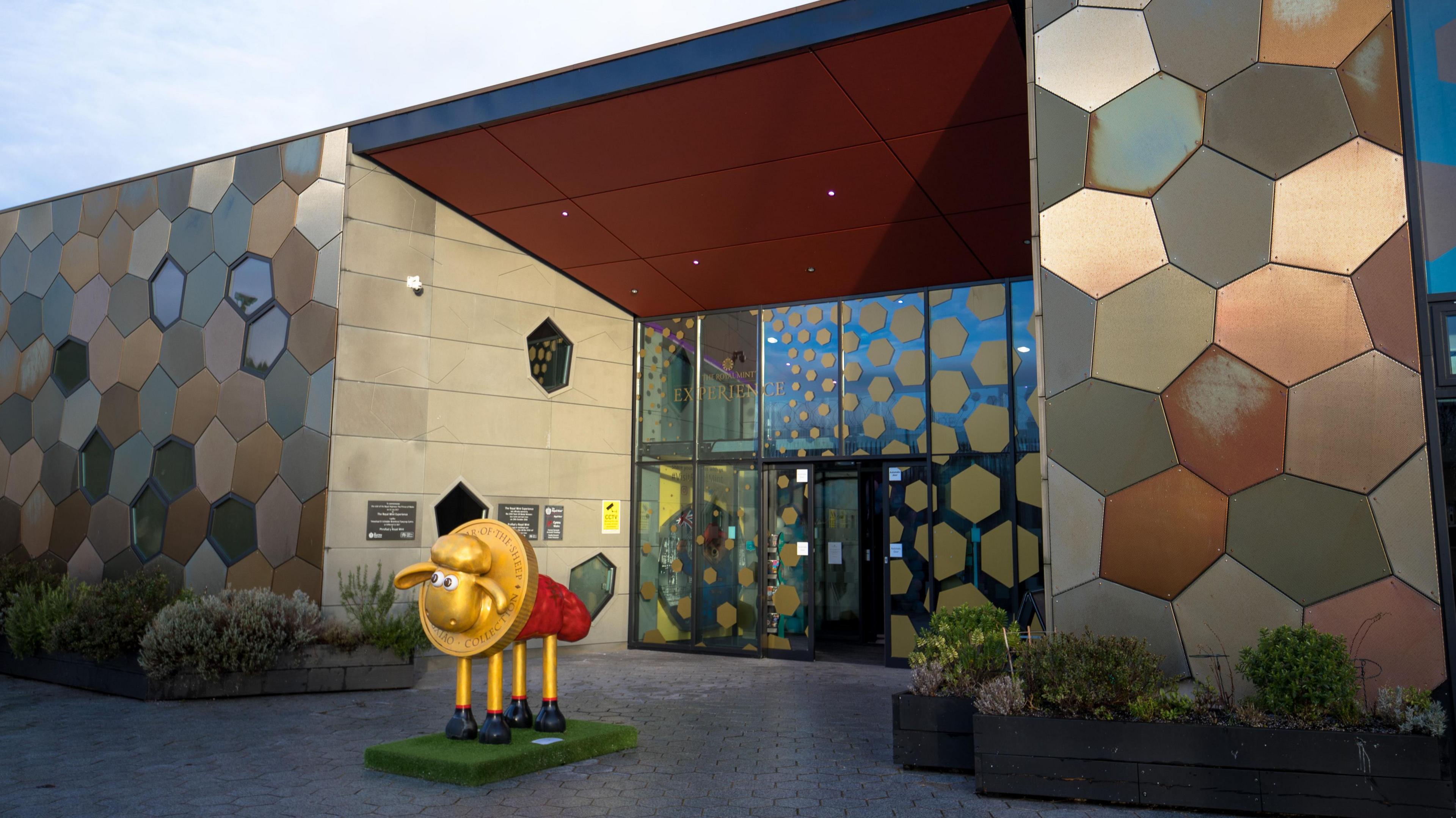 A boxy building clad in irregular hexagonal tiles. The motif is carried on into the glasswork of the entrance. In the courtyard there is a sculpture of Shaun the Sheep with its head protruding though the centre of a gold coin.