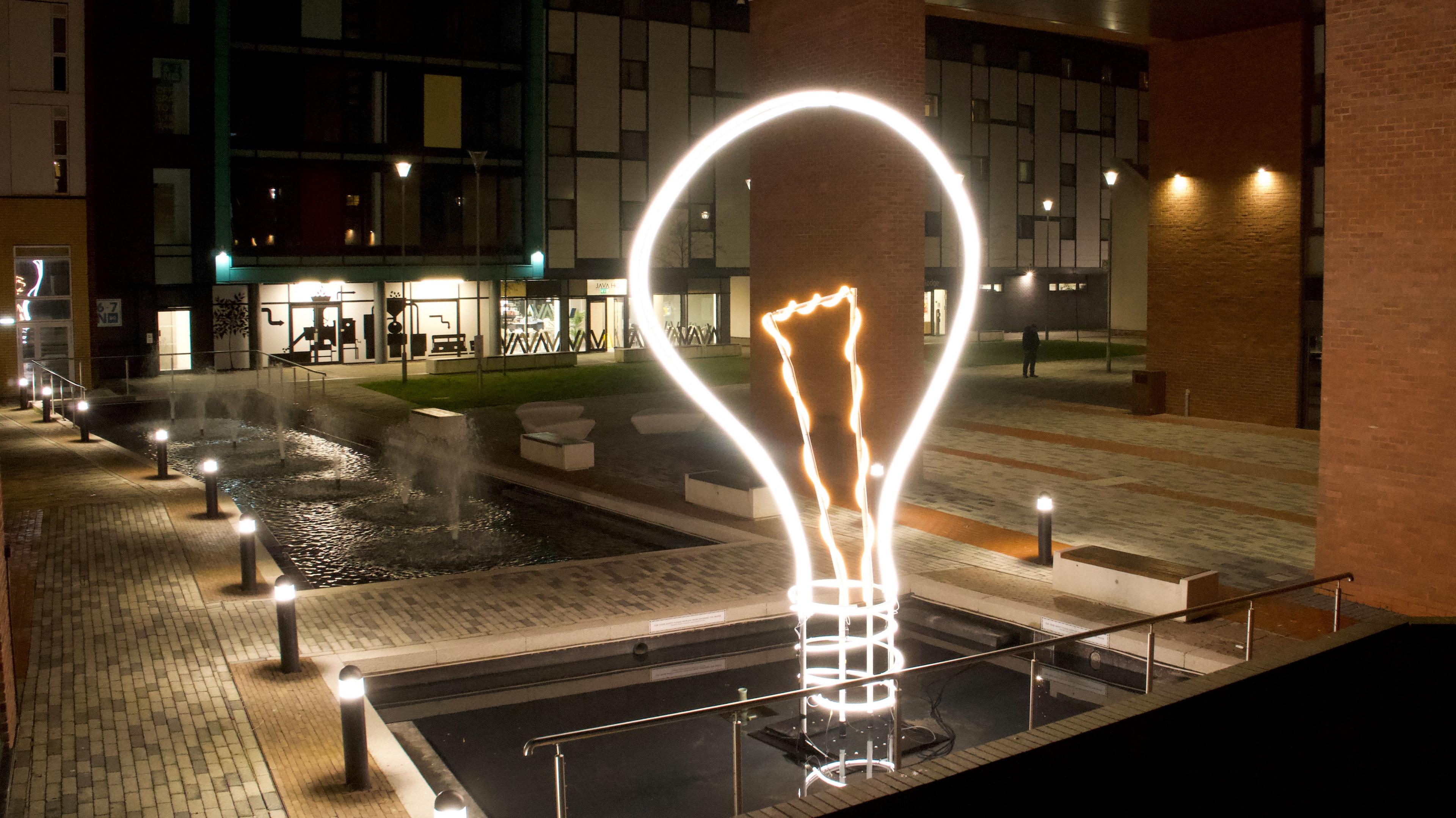 An outsize illuminated light bulb next to water fountains in a modern city centre square 