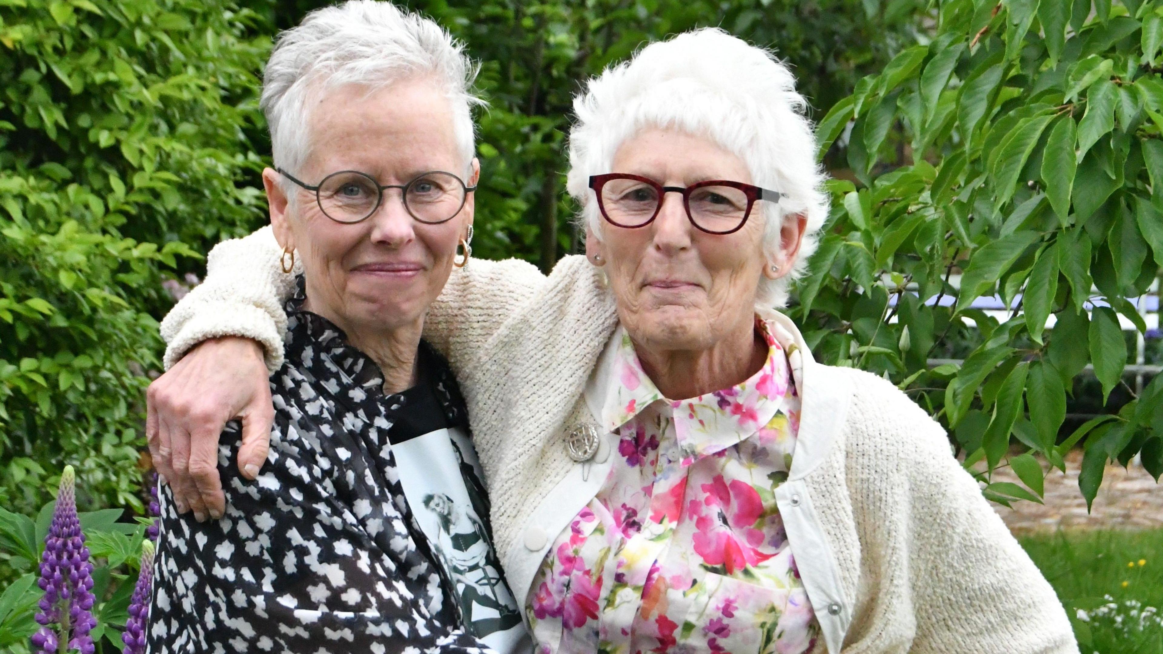 Anita and Steph, standing outside in a garden, with plants and trees. They are both smiling and looking straight at the camera. They both have glasses. Anita is wearing and black and white top, Steph, a floral top with a cream cardigan. 