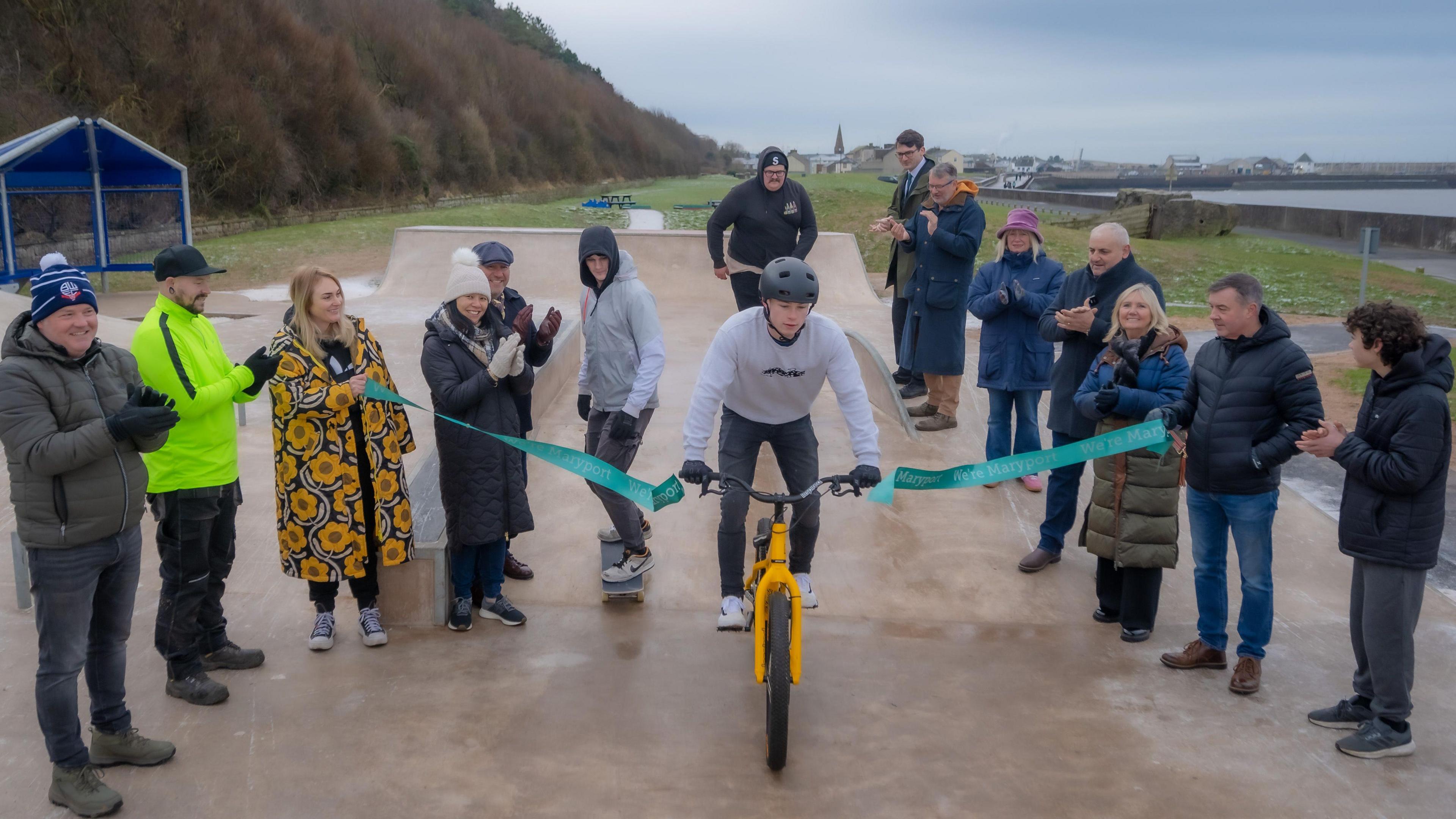 A young man cuts the ribbon by riding a BMX through it. He is surrounded by people clapping. In the background Maryport harbour is visible.