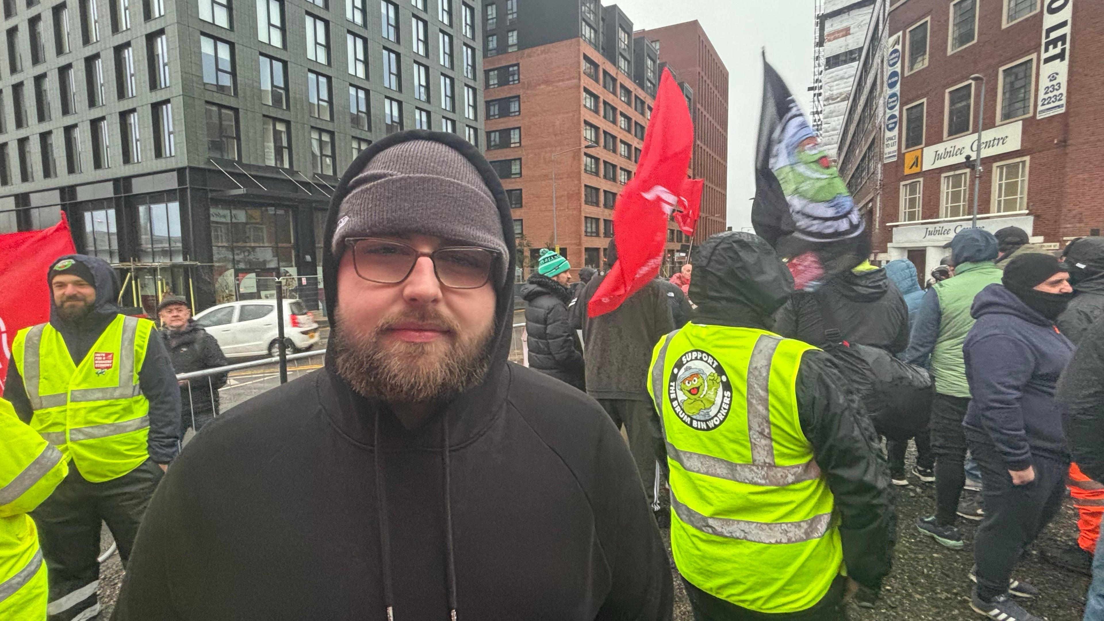 Agency worker Luke Graham wears a black hoodie with the hood up. He also has a grey woolly hat and glasses. Other workers can be seen behind him wearing bright yellow hi-vis jackets. Some are carrying flags.